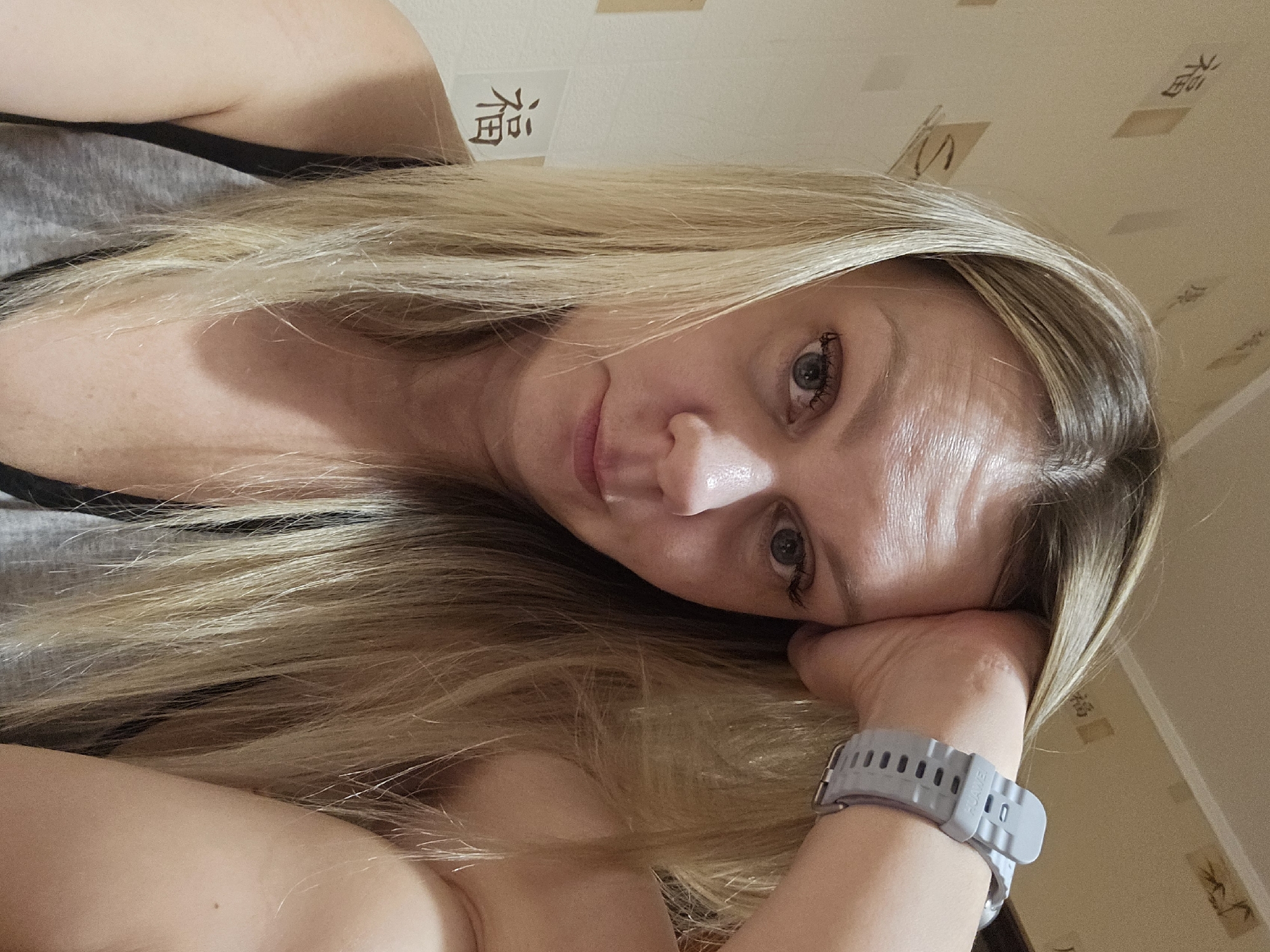 A woman with blonde hair is resting her head on her hand. She is wearing a gray watch. A black strap is visible under her gray top. A sign with Chinese characters is on the wall behind her. The wall has a light brown patterned wallpaper. The image does not provide information to confirm the location.
