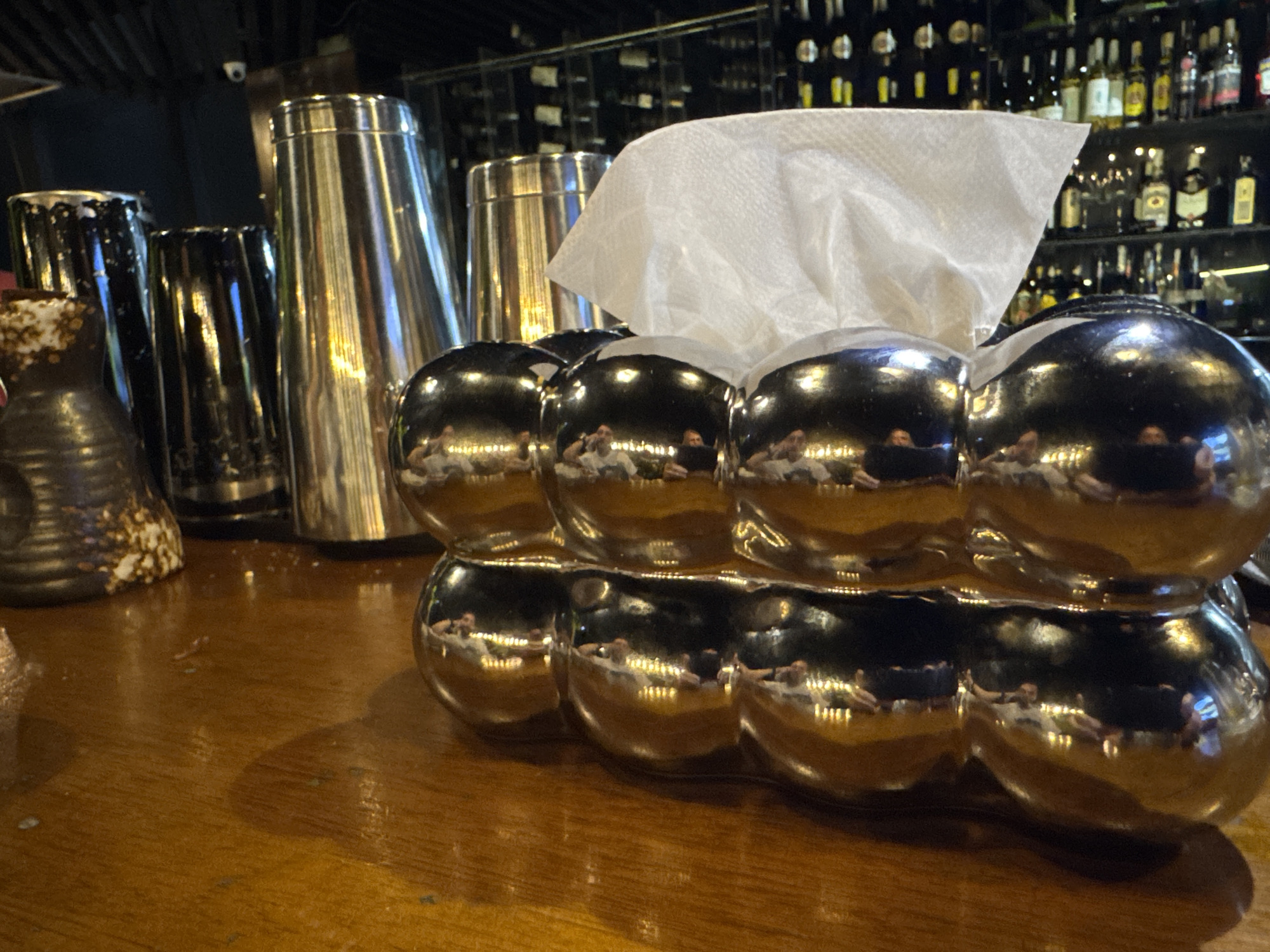 This image captures a close-up view of a wooden bar counter in what appears to be an evening or nighttime setting. The primary focus is a highly reflective, metallic tissue box with a distinctive bubbly, segmented design, positioned prominently in the foreground on the right side of the counter. White tissues are seen emerging from its top opening.
The polished surface of the tissue box, along with other metallic bar tools like cocktail shakers and containers situated behind it, creates clear reflections of the surrounding environment. Multiple human figures, appearing to be 3-4 individuals, are distinctly visible in these reflections, seated or standing, suggesting patrons or staff in the background. Their general shapes, light-colored clothing, and the presence of ambient light sources (possibly overhead lights) are noticeable within the reflections.
To the left of the tissue box, several metallic bar shakers and other cylindrical bar utensils are arranged. Further left, a small, dark-colored ceramic or pottery vase with white speckled details is visible. The wooden counter shows some signs of wear and minor smudges.
In the background, shelves are densely packed with numerous bottles of various shapes and sizes, strongly indicating an alcoholic beverage collection typical of a bar or lounge. The overall lighting is dim but sufficient to highlight the reflective surfaces and the objects on the counter. No specific activity or event is explicitly taking place, and no legible text is discernible on any bottles or labels. The scene offers a glimpse into a commercial drinking establishment.