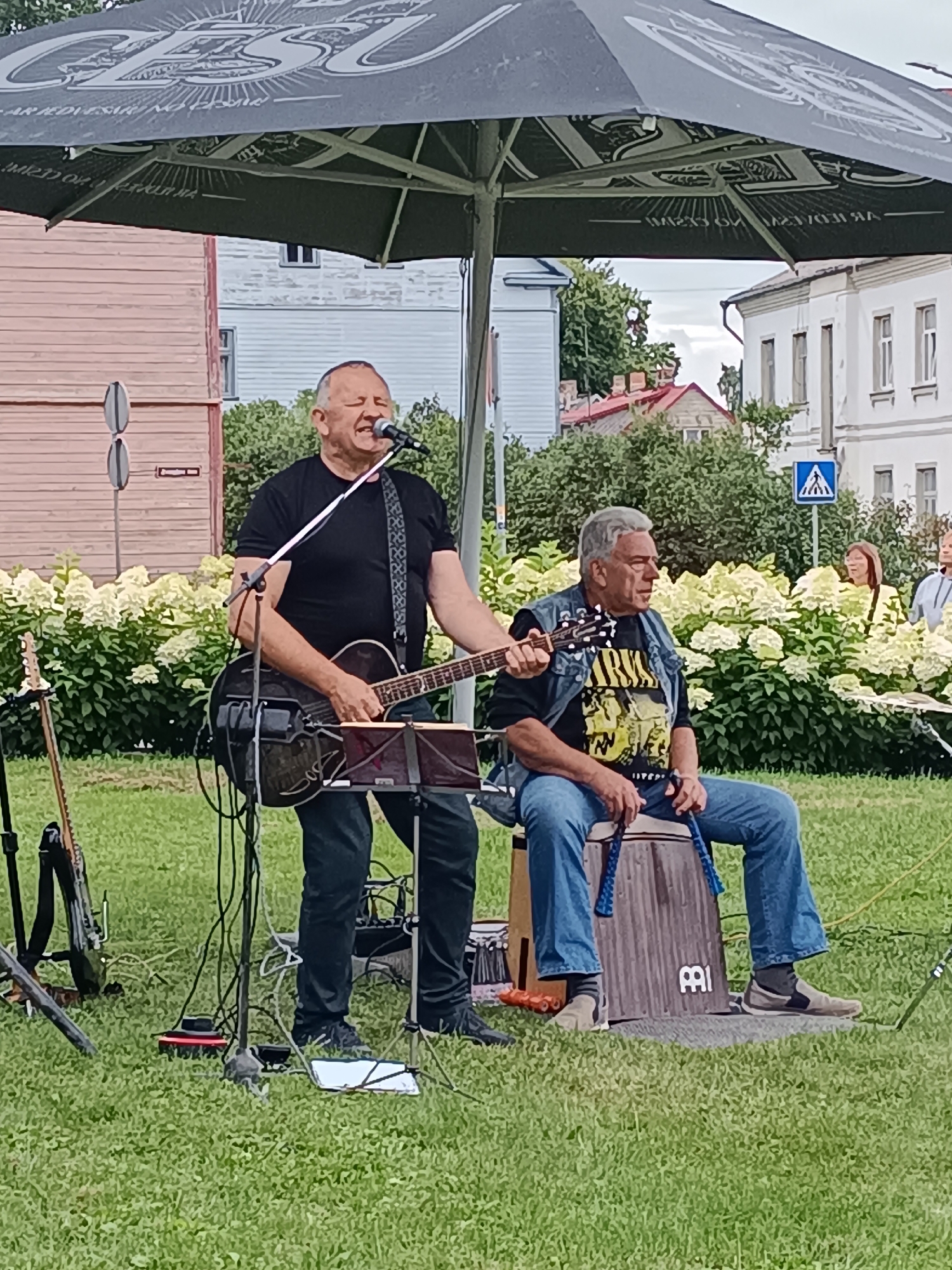 Two men are performing music outdoors under a large umbrella. The man on the left is singing and playing an acoustic guitar. He is wearing a black t-shirt and black pants. The man on the right is seated and playing a cajon drum. He is wearing a denim vest over a black t-shirt with yellow graphics and blue jeans. A third person, a woman, is visible in the background to the right, wearing a yellow top. The performance is taking place on a grassy area with a backdrop of flowering bushes and buildings. The umbrella has a logo that appears to be for "CĒSU" beer, suggesting the event may be sponsored by the brewery. The scene is consistent with an outdoor event or festival in the city of Aizpute, Latvia.