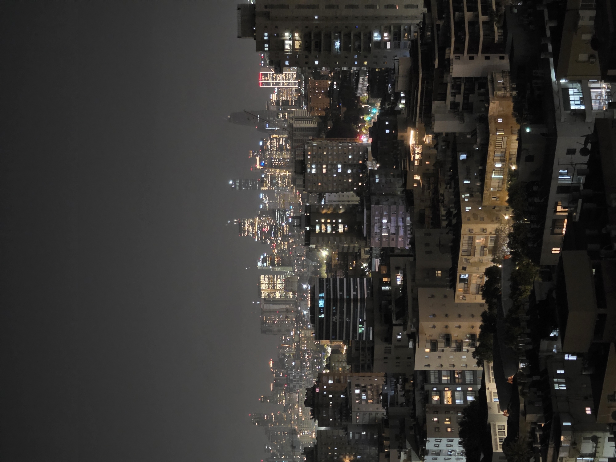 This elevated night view captures a sprawling urban landscape, likely Bat Yam, Israel, stretching across the horizon under a dark, possibly hazy sky. The scene is dominated by a multitude of buildings, all illuminated from within, creating a vibrant tapestry of light against the darkness.
In the immediate foreground and extending into the mid-ground on the right side of the image, various residential and commercial structures are visible. These buildings exhibit a range of architectural styles and heights, with countless windows casting warm, artificial light. Some buildings show balconies and air conditioning units, typical of an urban environment. Pockets of dark foliage from trees are interspersed among these structures.
Towards the left and extending into the distant background, the city transforms into a dense cluster of high-rise buildings and skyscrapers. The skyline twinkles with countless points of light from offices and apartments, gradually fading into a soft glow as they recede into the hazy distance. The diffused light suggests either atmospheric haze or perhaps distant mist. Construction cranes are visible among some of the taller buildings, indicating ongoing development within the city.
The overall impression is that of a bustling metropolis at rest, with its internal life illuminated for all to see. The time of day is clearly night, and the weather appears calm, with a slightly overcast or foggy sky diffusing the distant lights. There are no people visible, nor is there any discernible text within the frame. The image emphasizes the architectural grandeur and the constant energy of a modern city after dark.