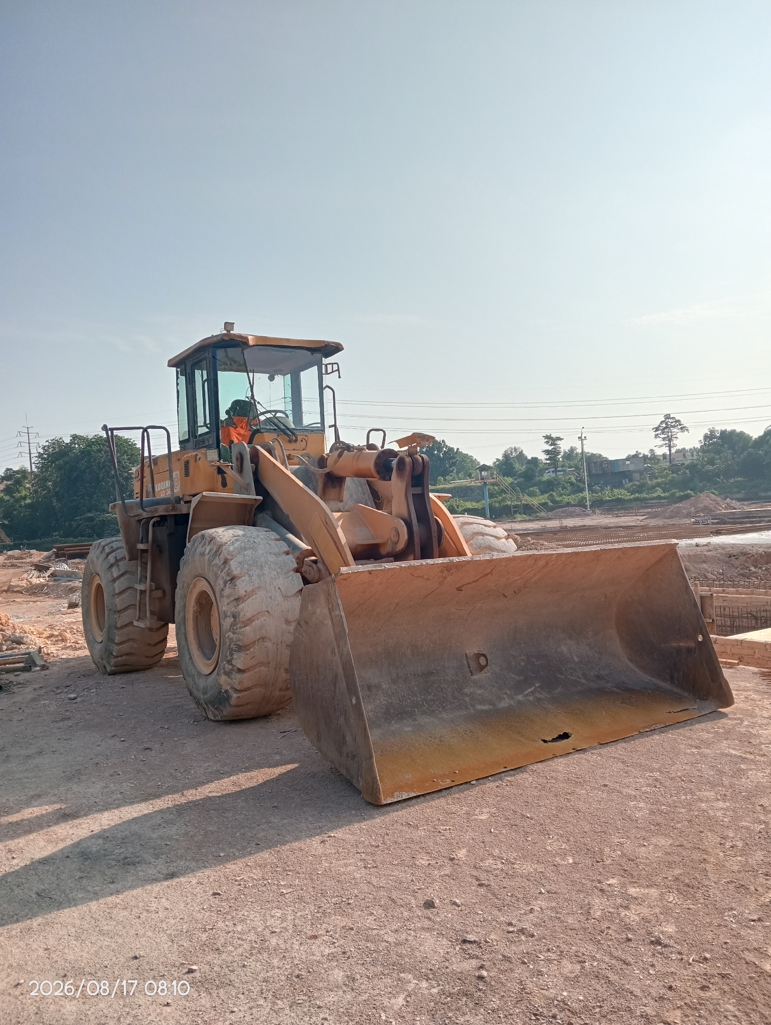 An orange front-end loader sits on a dirt construction site under a clear sky. The machine, labeled "LUIGINI LO DE," has large tires and a wide bucket. In the background, trees and power lines are visible, along with unfinished concrete structures. The timestamp "2026/08/17 08:10" is present in the lower left corner of the image. The scene appears to be a construction zone in Batam, Indonesia.