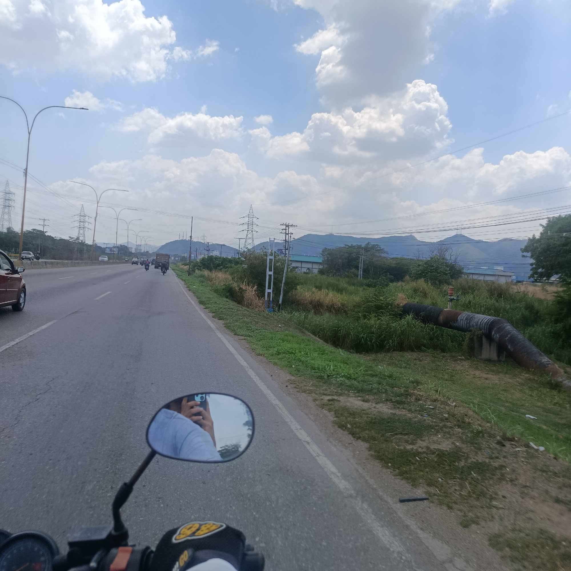 A view from a motorcycle riding on a highway in Los Guayos, Venezuela, shows a multi-lane road with light-colored asphalt under a partly cloudy sky. Power lines and towers run parallel to the road. Other vehicles are visible in the distance. To the right of the road is vegetation and a large exposed pipe. The motorcycle's side mirror reflects the rider taking a photograph with a mobile phone.