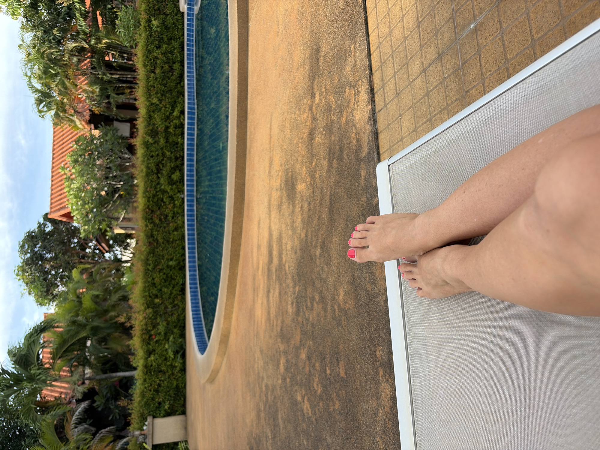 In Baan Mae Nam, Thailand, a pair of bare feet with pink toenails rests on a light grey mesh lounge chair. The feet are crossed at the ankles. In the background, a curved swimming pool with blue tiles is visible, surrounded by a tan paved deck. Beyond the pool, a well-maintained green hedge borders a pathway. Further in the background, traditional Thai-style buildings with orange tiled roofs are nestled among palm trees and lush green foliage under a partly cloudy sky.