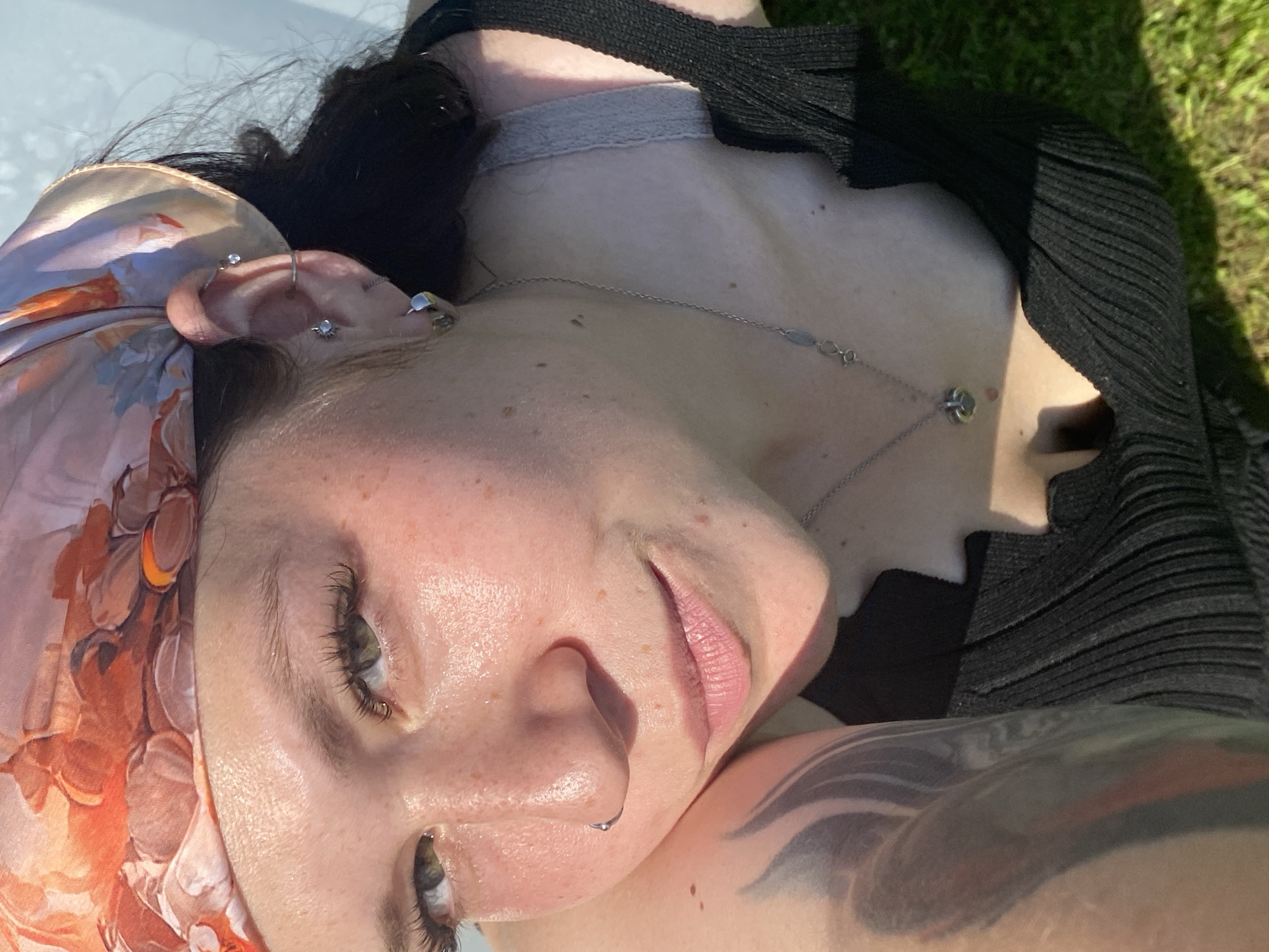 The image shows a close-up, overhead view of a young woman lying on green grass, wearing a black ribbed tank top and a patterned headscarf. She has freckles on her face and arms, and several piercings in her ears, along with a nose ring. A silver necklace with a heart-shaped pendant is visible around her neck. A large tattoo is present on her left arm. The background appears to be a light blue surface, possibly a wall or a pool edge, suggesting an outdoor setting. The overall lighting indicates it is daytime, with sunlight casting shadows and highlighting the textures of her skin and clothing. The location is not specified within the image itself, and therefore cannot be determined to be in the city of Фоки, Russia.