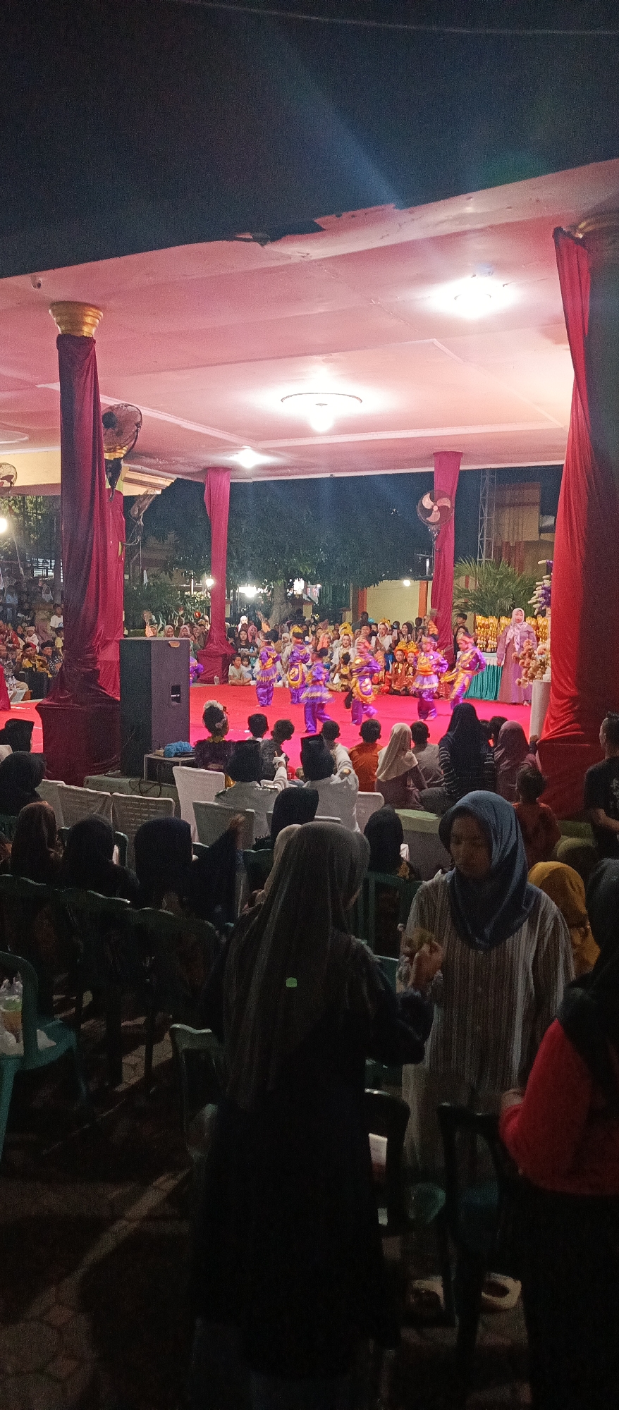 An evening outdoor cultural performance is taking place on a raised red carpeted stage under a pink illuminated canopy in Arjasa, Indonesia. A group of children dressed in colorful purple and gold traditional costumes are performing a dance center stage. The audience is seated on chairs in the foreground, with many wearing headscarves. The stage is flanked by large maroon velvet curtains draped over pillars. A large black speaker is positioned to the left of the stage. The environment appears festive, with decorative elements and lighting contributing to the atmosphere.