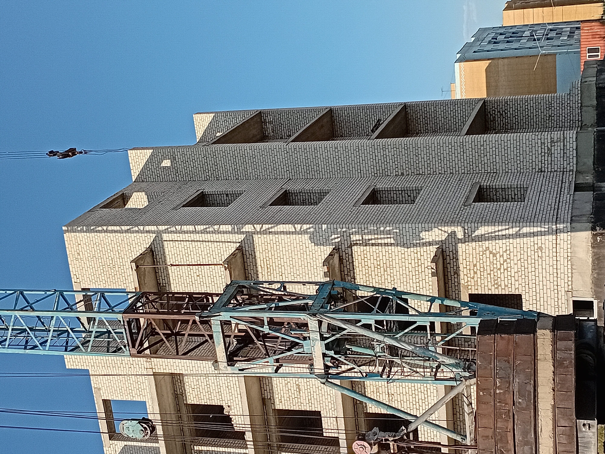 The image shows a construction site in the city of Semey, Kazakhstan. A large tower crane is positioned next to a multi-story building under construction, which is made of light-colored bricks and has unfinished balconies and window openings. The sky is clear and blue. In the foreground, the counterweights of the crane are visible, consisting of stacked metal plates. Further in the background, other buildings, also appearing to be residential, are visible. The scene depicts a typical urban construction environment.