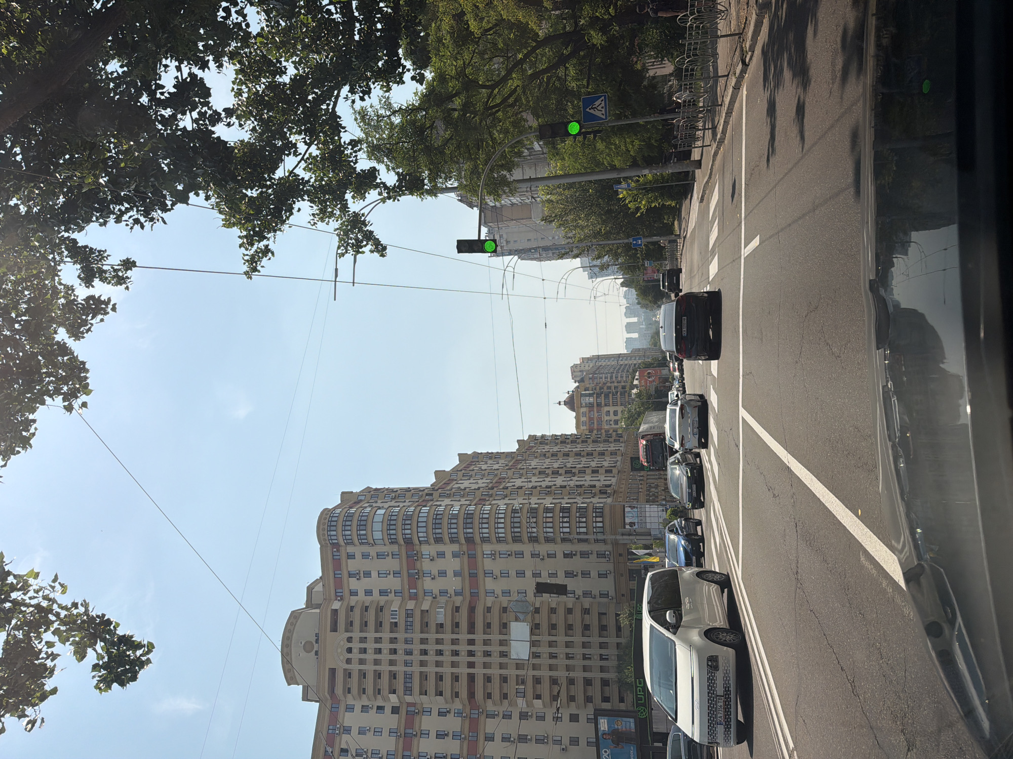 The image shows a street scene in Kyiv, Ukraine, with a focus on traffic and urban architecture. The perspective is from inside a vehicle, looking forward and slightly to the left. Several cars are visible in lanes on the right side of the road, with a traffic light showing a green signal in the distance. Tall, modern apartment buildings line the left side of the street, featuring a distinctive curved facade and numerous windows. Lush green trees with dense foliage are prominent, casting shadows on the road and sidewalk, suggesting it is daytime. The sky is clear and blue. On the sidewalk, bicycle racks are visible, and a pedestrian crossing is marked on the asphalt. Some billboards or advertisements can be seen on the side of the road. The overall impression is a bustling urban environment on a sunny day. There is visible text on a license plate of a white car that appears to be in Cyrillic script.