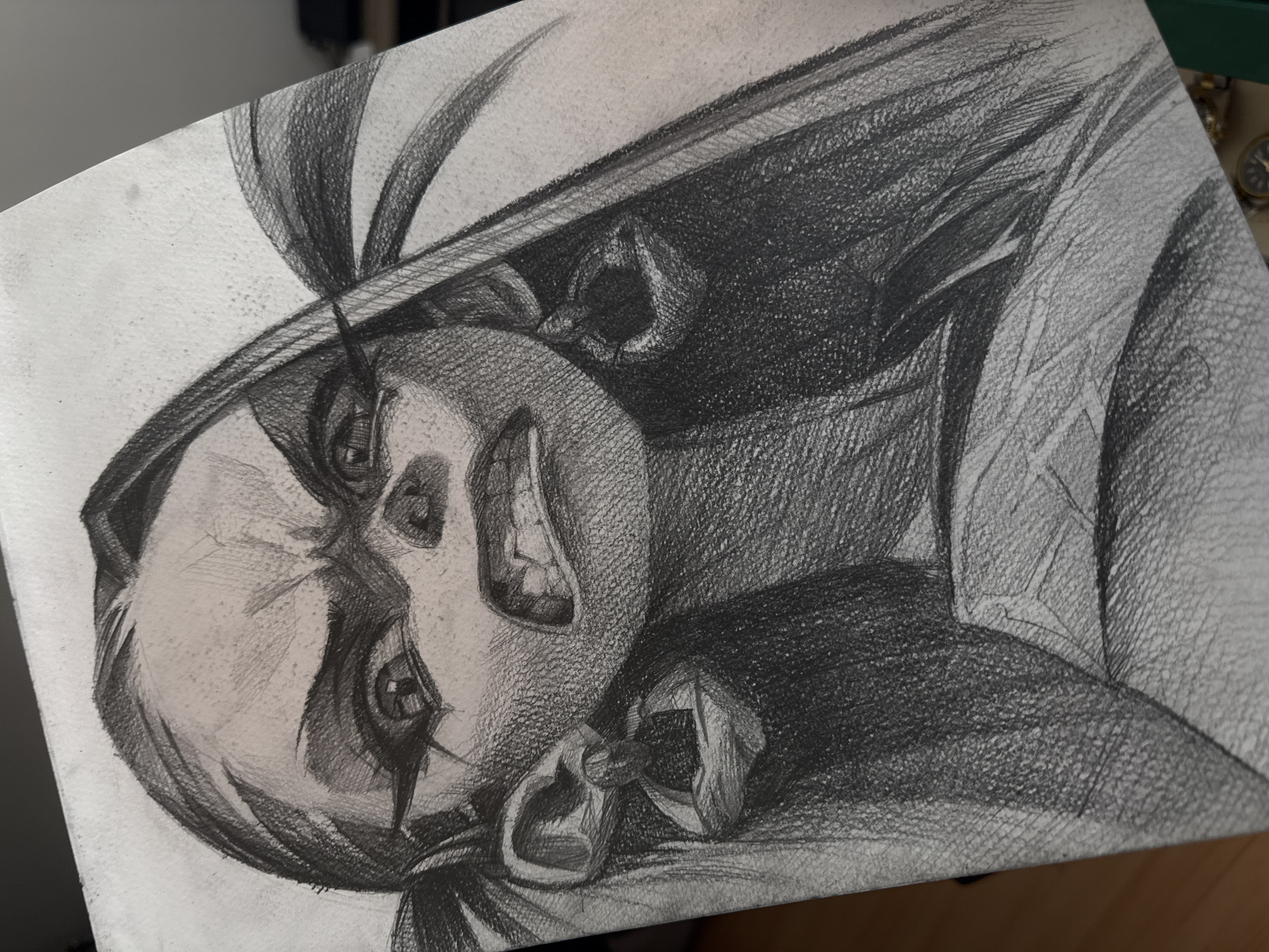 The image shows a pencil drawing of a stylized character. The character has a large, round head with exaggerated features, including large eyes, a prominent nose, and an open mouth with visible teeth. The character is wearing a hat with what appears to be a feather or plume. Dark shading creates a sense of depth and dimension, particularly around the hat and the back of the character. The drawing is on a sheet of paper. The background is out of focus. The image is rotated counterclockwise. The location is not discernible.