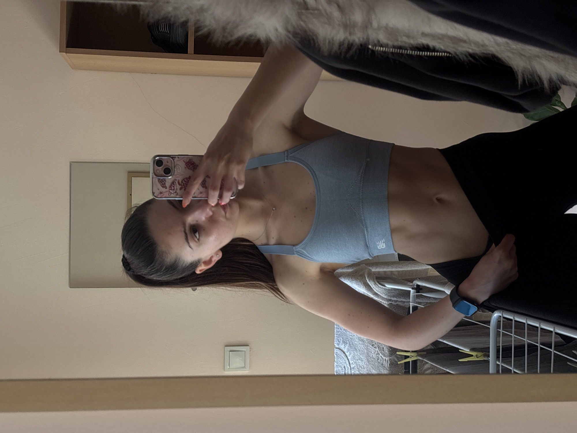 A young woman is captured in a mirror selfie, standing upright and holding an iPhone with a patterned case (featuring what appear to be turtles) in her right hand, aimed at her reflection. She has long, dark hair pulled back and looks towards the phone's camera with a neutral expression.
She is dressed in a light blue ribbed sports bra, which has a white "NB" (New Balance) logo on the left strap area, and black leggings. A delicate silver necklace adorns her neck, and a blue-banded Apple Watch is worn on her left wrist. Her fingernails are painted a light orange or coral color.
The setting appears to be an indoor room, likely a home, with various objects reflected in the mirror. In the upper left of the background, a wooden shelf or cabinet is visible, and above the woman's torso, a fluffy, possibly white, garment hangs. A white light switch is mounted on the wall in the lower left. To the lower right, a white textured towel is draped over an object, and part of a white metal drying rack with yellow clothes pegs can be seen, indicating a domestic environment. The woman exhibits a fit physique, with visible abdominal muscles. The lighting is uniform, typical of an indoor shot.