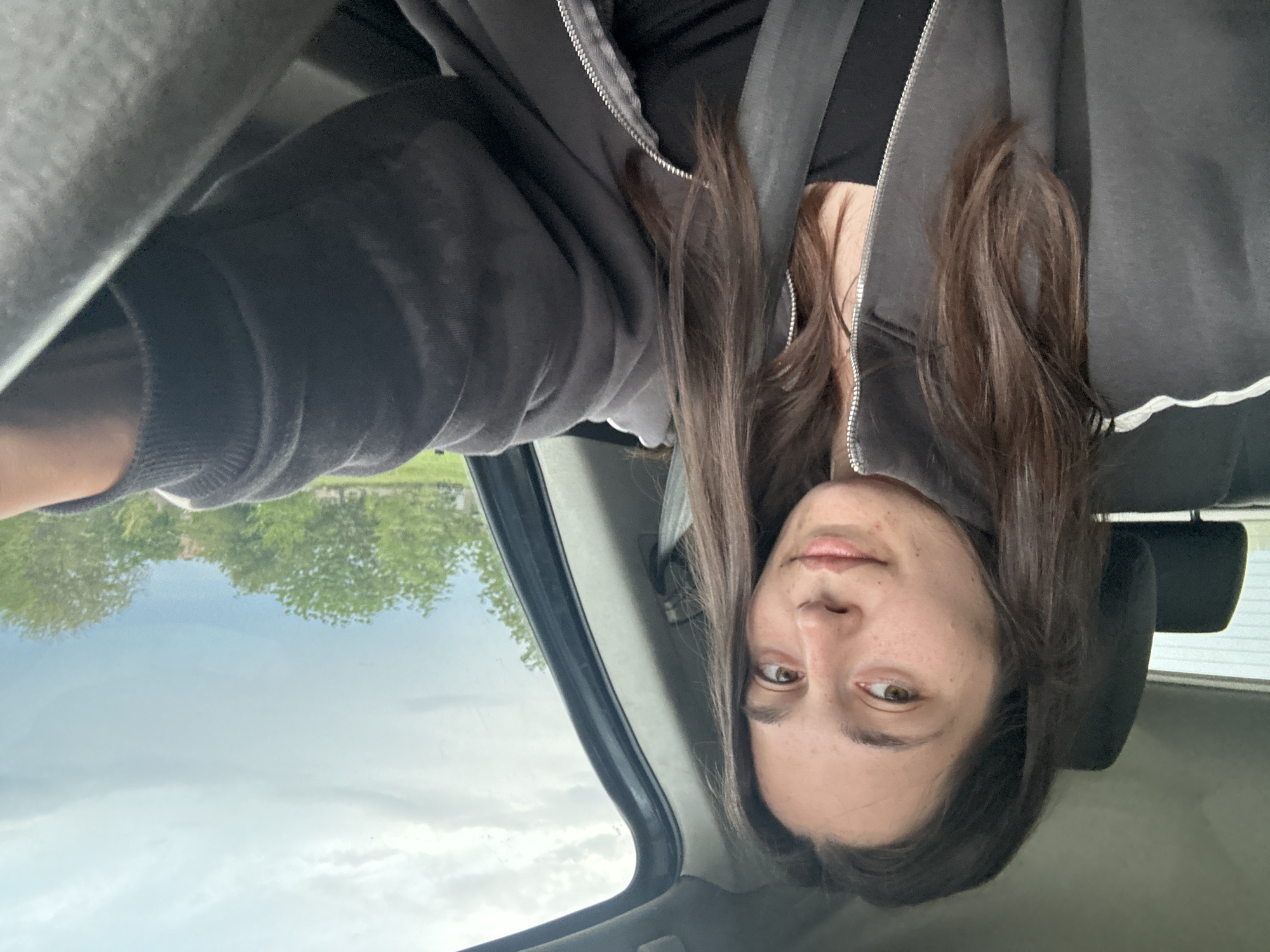 A woman is sitting in a car, wearing a black top, a dark grey zipped-up hooded sweatshirt, and a seatbelt. The background shows a cloudy sky, trees, and buildings that suggest an outdoor environment. It is difficult to establish the precise location within Resen, Bulgaria, based solely on this image.