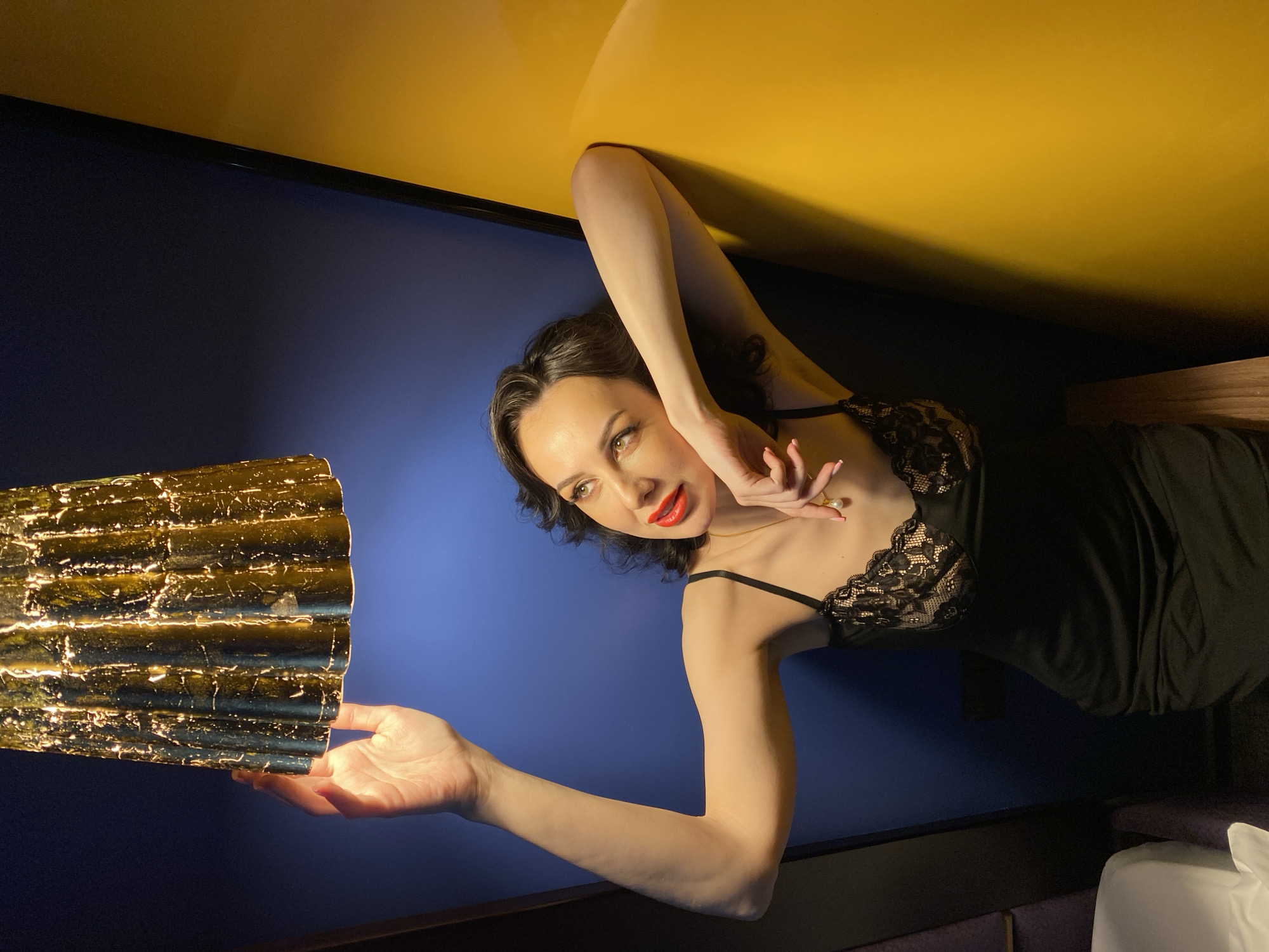 A female individual is lying down. She has black hair and is wearing a black dress with lace details. She is positioned next to a gold, textured lamp, her hand near the lamp's light source. The background features a blue wall and a yellow ceiling. There is no discernible information to identify the location as Yekaterinburg, Russia.