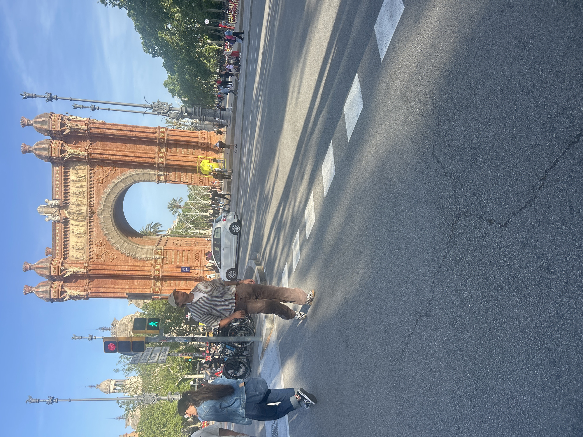 The image depicts a street scene in Barcelona, Spain, featuring the Arc de Triomf as the prominent architectural element. The Arc, constructed of reddish-brown brick, stands on the left side of the frame. A busy pedestrian crosswalk is visible in the foreground of the image. The crosswalk extends across the street, marked by white rectangular lines. Several people are captured mid-crossing, including a man in a striped shirt and brown pants, and a woman wearing a blue denim jacket and jeans. The pedestrian traffic light displays a green signal for crossing. A line of parked bicycles are behind the crossing pedestrians. A street with a few cars separates the street. Behind, there are multiple people crossing the street. A blue sky is visible above the buildings.