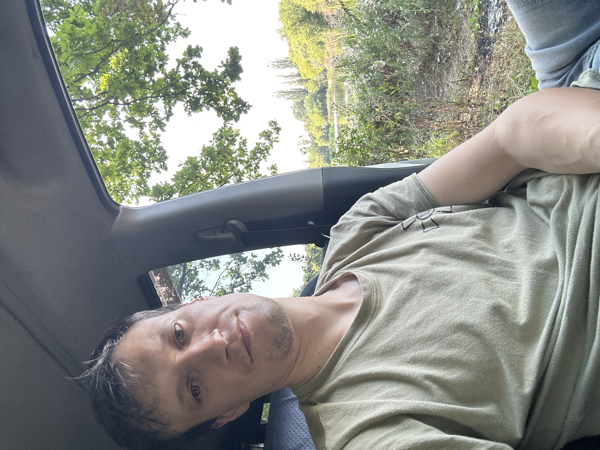 A man with short brown hair and light-colored eyes is seated in the passenger seat of a vehicle. He is wearing a light olive green t-shirt and blue jeans. His left arm rests on his lap, and his right arm is visible bent at the elbow. The man is looking towards the camera with a neutral expression.
The interior of the vehicle is visible, including the headliner, a portion of the driver's seat, and a seatbelt buckle. Through the open passenger side window, a natural landscape is visible. This includes a body of water, trees, and vegetation. The foliage appears green and lush, suggesting a temperate climate. The sky is bright and appears to be daytime.
The location is identified as Makiv, Ukraine. The scene depicts a moment of rest or travel through a rural or semi-rural area.