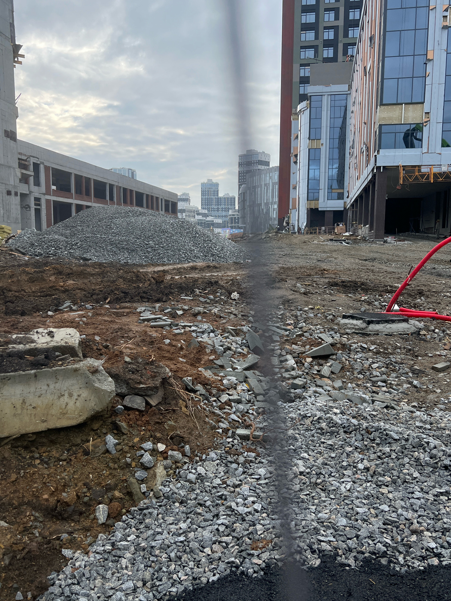 The image depicts a construction site in the city of Yekaterinburg, Russia. In the foreground, there is a pile of gravel and debris, with some concrete fragments. A trench or channel filled with dark material runs through the gravel. In the middle ground, a large mound of gravel is situated in front of a concrete structure with brick infill. Further back, modern multi-story buildings with glass facades are visible, suggesting an urban environment. One building, in particular, appears to be under construction, with scaffolding and exposed structural elements. A red pipe is visible in the right midground, emerging from a manhole cover. The sky is overcast with clouds. The overall scene is one of ongoing construction activity in a developing urban area.