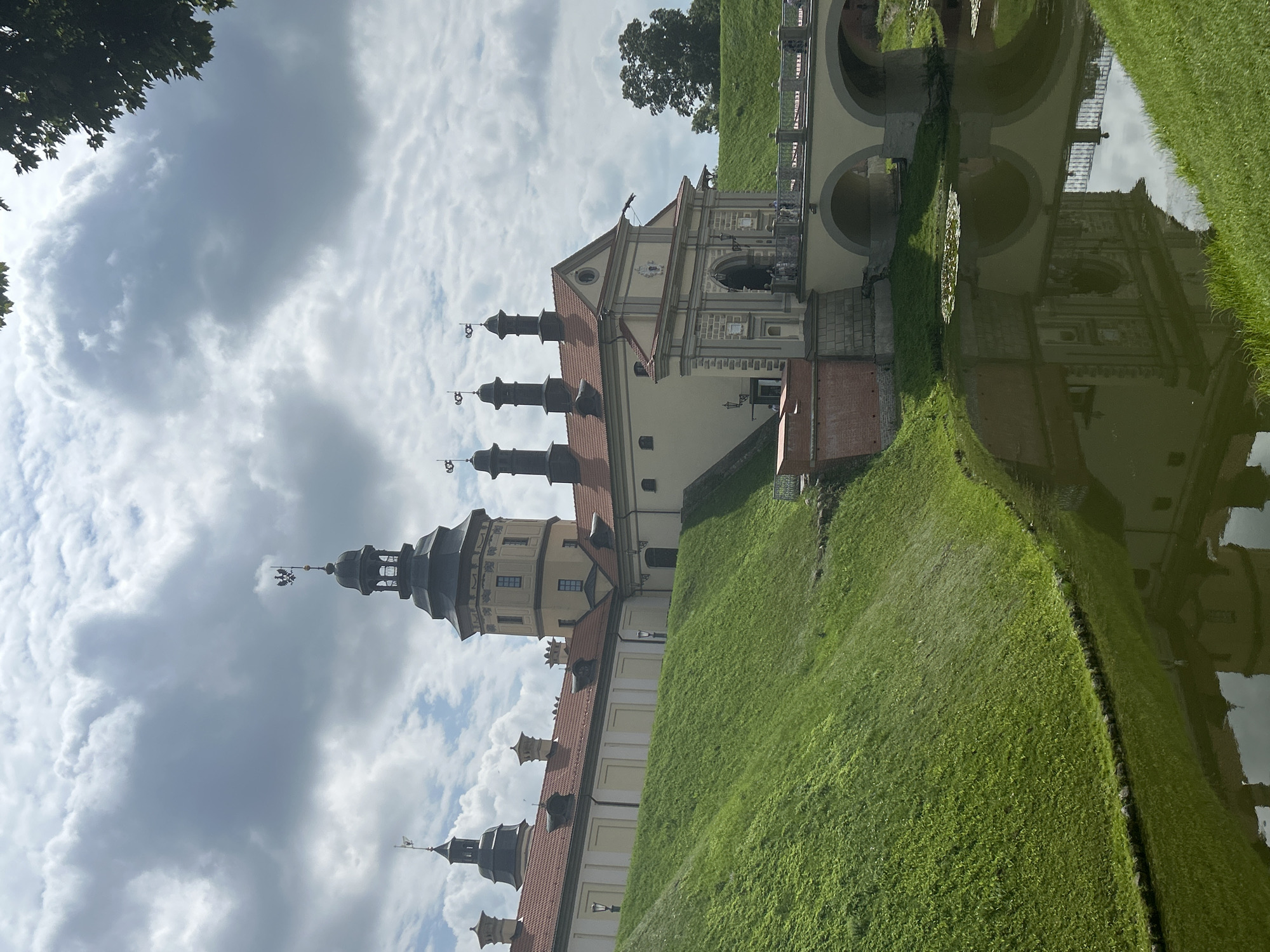 The image shows a view of Nesvizh Castle, a UNESCO World Heritage site located in Nesvizh, Belarus. The castle is a majestic structure with a large central tower topped with a dark spire and a weather vane. Several other smaller towers and buildings with red tile roofs are visible, along with a grand entrance gate. A stone bridge with arches spans a moat filled with green water, reflecting the castle's architecture. The castle is surrounded by lush green grassy slopes. The sky above is partly cloudy, with dramatic white clouds against a blue sky. A few people are visible on the bridge and near the castle entrance. The overall scene conveys a sense of historical grandeur and natural beauty.
