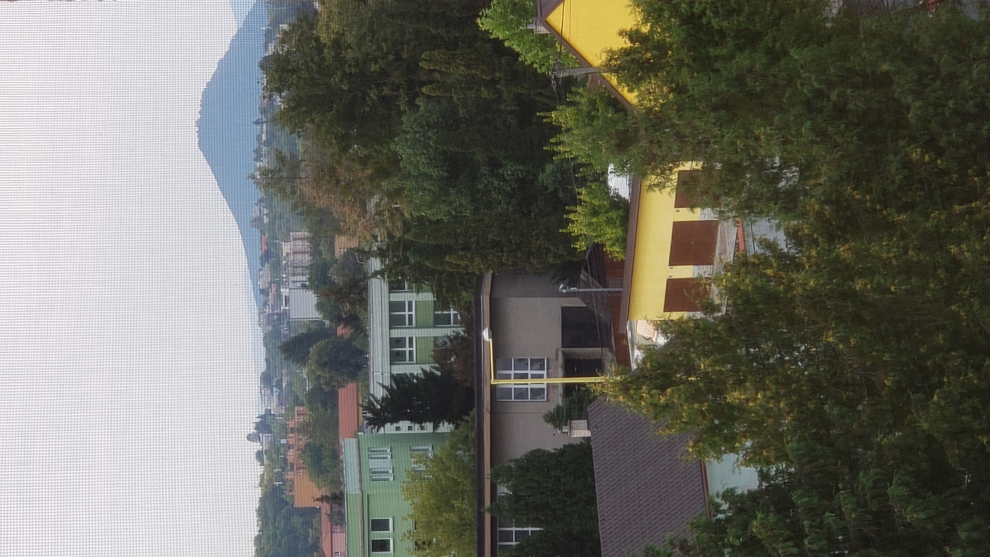 The image displays a residential area with several buildings and abundant trees, set against a hazy sky and a distant mountain. In the foreground, a cluster of tall evergreen trees partially obscures a view of buildings. Behind these trees, a low, grey building with a prominent light pole in front of it is visible, featuring large windows and dark garage doors. To the right of this building, a portion of a bright yellow structure with a brown garage door is seen. Further back, there are multi-story buildings, including one with a green facade and white-framed windows, and another with a beige facade and reddish-brown roof. In the distance, a prominent, cone-shaped mountain dominates the horizon, with a small settlement or structures visible on its peak. The overall impression is that of a somewhat overcast day in a suburban or urban environment. While no people are directly visible, the presence of residential buildings and vehicles suggests human activity. The image appears to have been taken through a screen, indicated by a faint grid pattern overlaying the scene. The location is identified as Prešov, Slovakia, and the mountain in the background is likely the Zemplín Mountains.