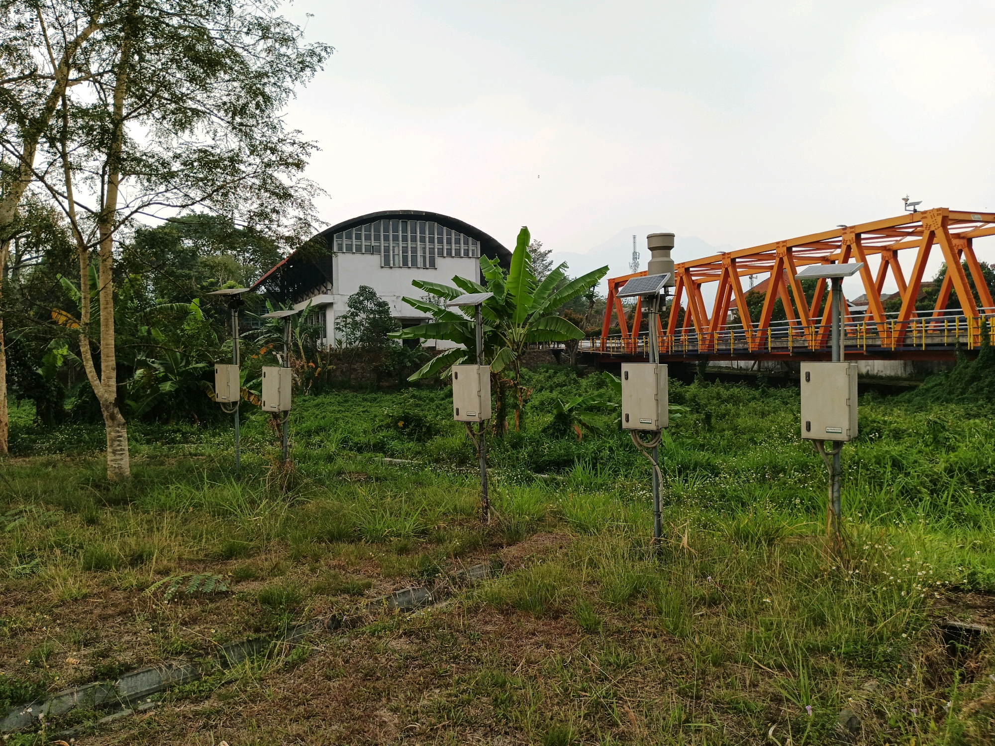 In the city of Bandung, Indonesia, a scene unfolds with a modern bridge and a large building adjacent to a grassy area. The bridge, painted a vibrant orange, spans a waterway and features yellow railings. On the bridge and near it, several solar panels are visible, suggesting a renewable energy source. A white building with a distinctive curved roof and numerous windows stands to the left of the bridge, surrounded by lush greenery, including banana trees. In the foreground, five small, gray posts with white boxes and solar panels are arranged in a line. These structures are in a field of tall grass and some dry vegetation. The overall impression is one of infrastructure integrated with nature in an urban or semi-urban setting. The sky is overcast, with a few distant mountains visible in the background. There are no people present in the image.