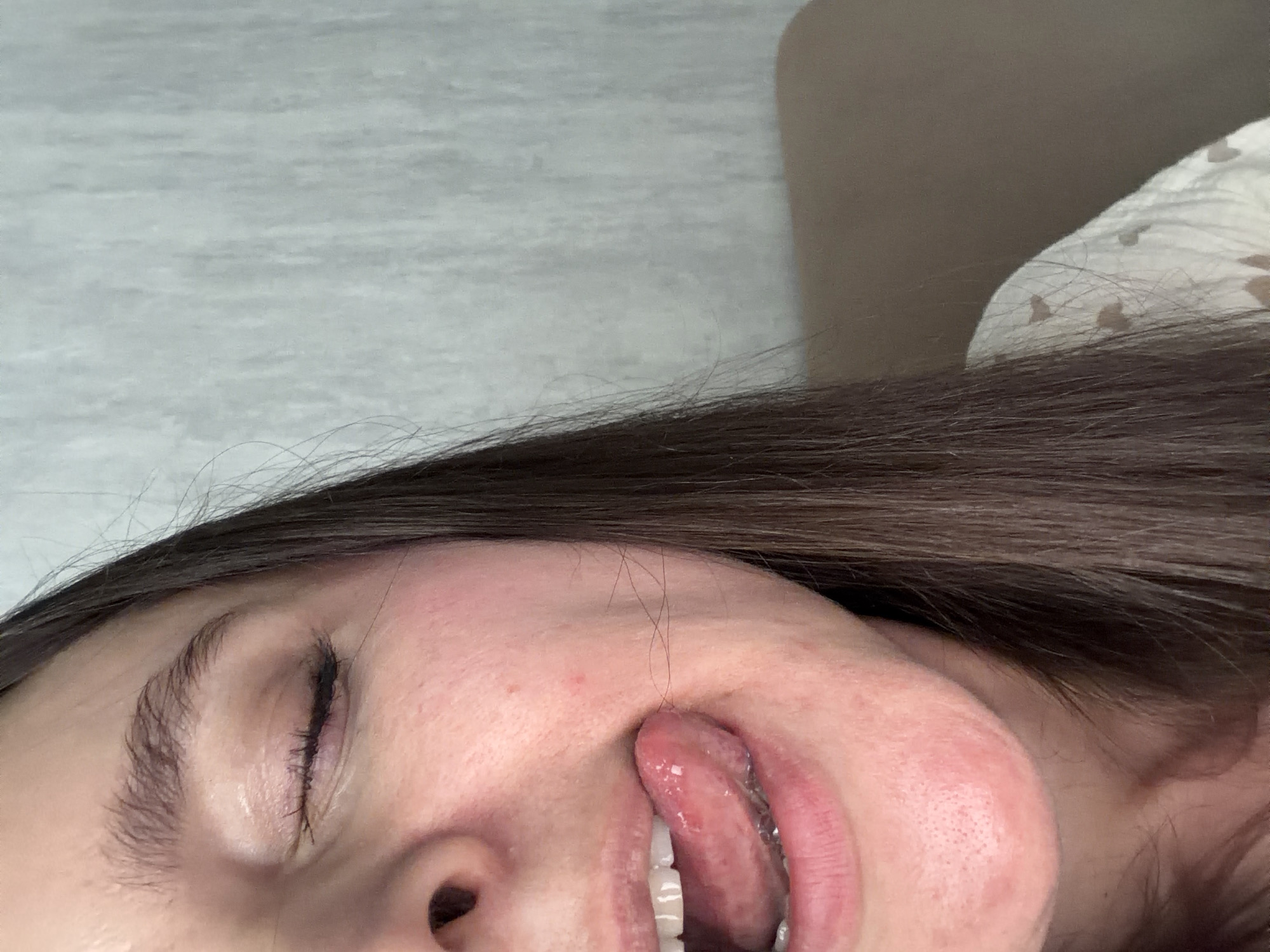 A close-up, eye-level, indoor shot shows the left side of a young woman's face and upper chest against a lightly textured, pale gray background. She has dark brown hair and fair skin. Her left eye is closed, with visible eyelashes and eyebrow. Her mouth is open, with her tongue sticking out and touching the left side of her upper lip. Her teeth are visible. Her chin area shows some reddening and visible pores. She is wearing a white top with a subtle pattern of small, light brown hearts. The setting is an indoor environment, possibly a home, with no specific landmarks to identify it as being in Shushary, Russia.