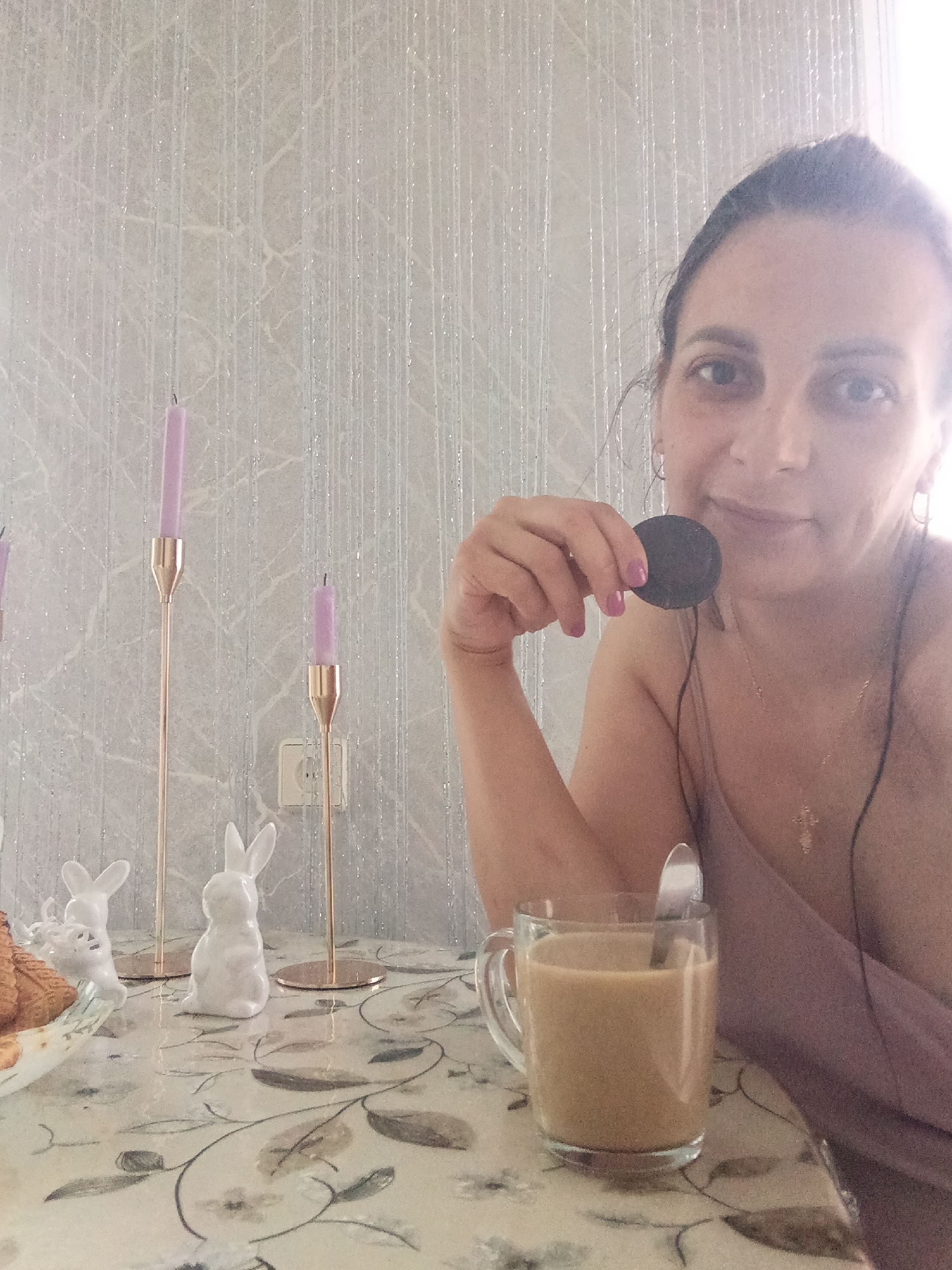 A woman is seated at a table in Andreyevo, Russia. She is holding a chocolate cookie in her right hand. A clear glass mug containing a light brown liquid, likely coffee or tea with milk, is on the table. A spoon is inside the mug. There is also a plate of pastries and two white ceramic rabbits on the table. Behind these objects are two tall, gold-colored candle holders with purple candles. The wallpaper is light-colored with vertical stripes and a subtle floral pattern. The table has a floral tablecloth. The woman is wearing a light purple top, a necklace with a cross, and small hoop earrings. She has her hair pulled back. An electrical outlet is visible in the background.
