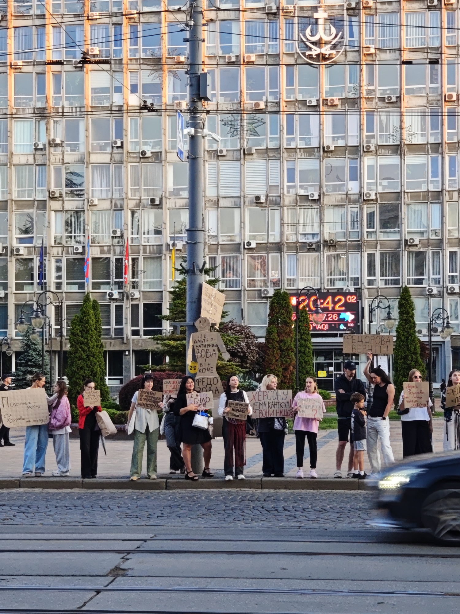 The image captures a group of people protesting in front of a large, modern building with many windows, possibly an administrative or governmental building. The building's facade is a pale concrete color, and the windows are evenly spaced. A large digital clock displaying the time "20:42" is visible on the building's exterior. The protest appears to be taking place on a paved street or plaza, with tram tracks visible in the foreground. Several individuals are holding signs with text, suggesting a political or social demonstration. The people are dressed in casual clothing, and some are standing, while others are holding their signs aloft. The overall atmosphere seems to be one of public assembly and expression of views. The presence of tram tracks and the style of the building suggest an urban setting in Eastern Europe, and given the context of ongoing events, it is plausible this location is in Vinnytsia, Ukraine.