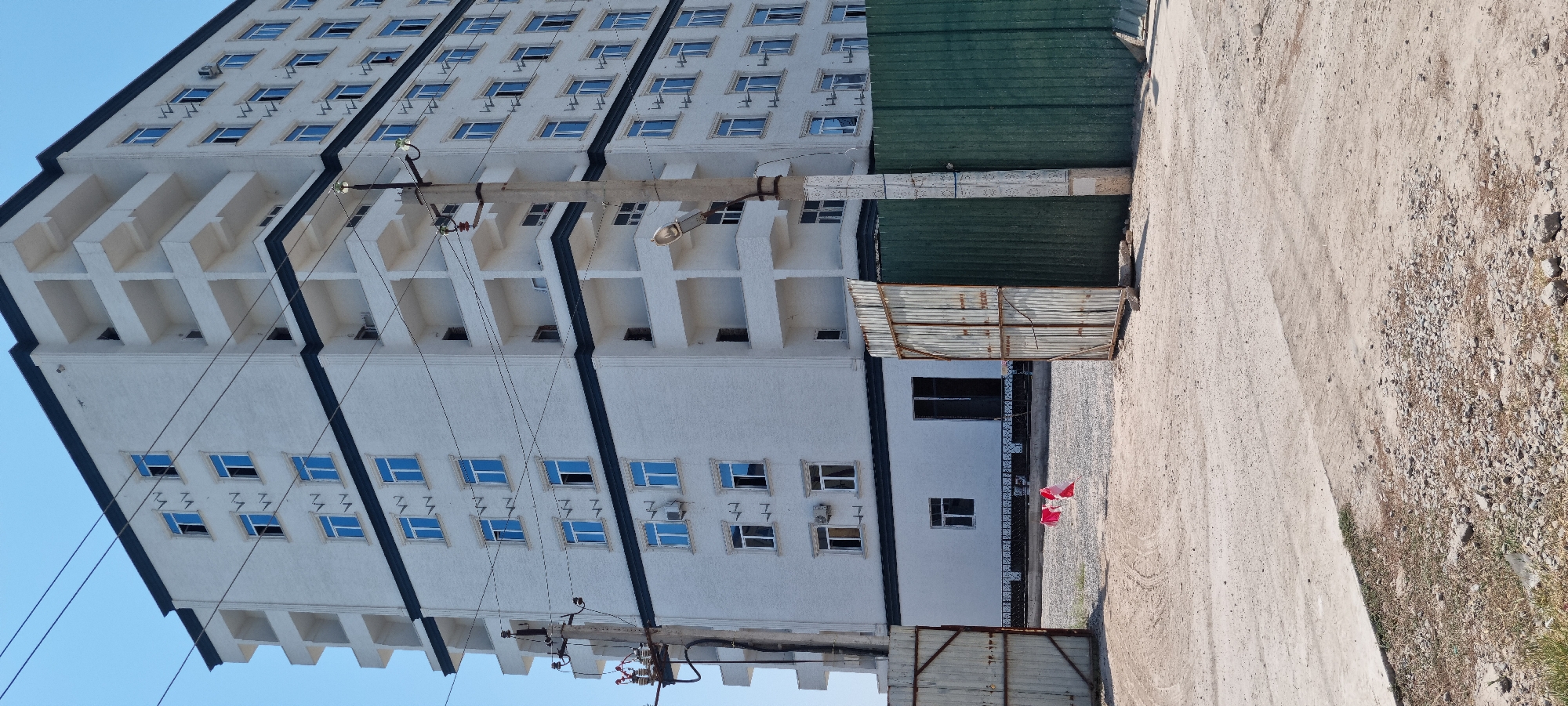 In the city of Ismoili Somoni, Tajikistan, a large, multi-story white building with dark trim and balconies stands under a clear blue sky. Power lines crisscross the frame in front of the building, supported by a utility pole. To the left of the building, a large open metal gate leads to a construction site with gravel and tire tracks on the ground. To the right of the building, a green corrugated metal fence encloses a portion of the site, with another rusty metal gate in front of it. A red and white marker lies on the ground near the center of the image, suggesting ongoing construction or work. The building appears to be a modern apartment complex or office building, possibly under construction or recently completed.