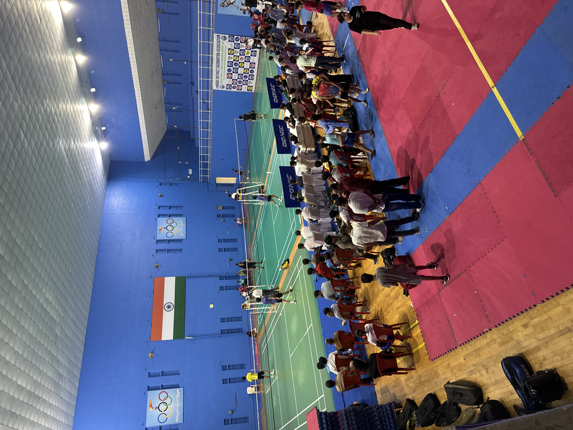 The image shows an indoor badminton court with a match in progress. Spectators are seated in the foreground, and players are on the court. The court is green with white lines, and the surrounding walls are blue. There are banners on the walls, including the Olympic rings and the Indian flag. The lighting in the venue appears to be artificial, with several lights visible on the ceiling. The overall scene depicts a sporting event in a sports facility.