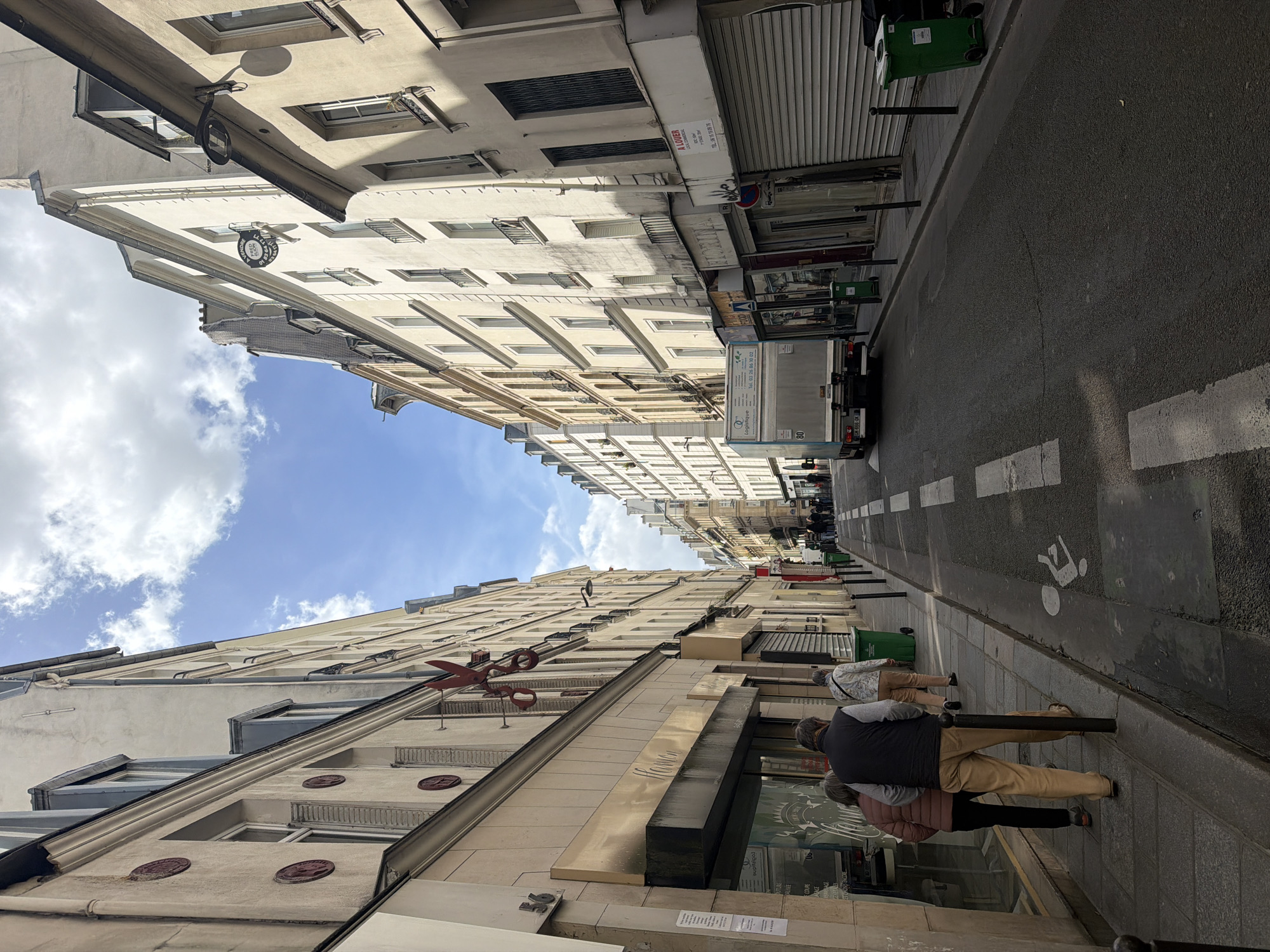 This media file captures a bright, sunny street scene in an urban environment, likely Paris, looking down a narrow street flanked by tall, traditional Haussmannian-style buildings. The image is taken from a low angle, emphasizing the height of the buildings and the clear blue sky with scattered white clouds visible above.
In the foreground on the left, a multi-story building features ornate window detailing and balconies. A large, decorative red sculpture resembling a pair of open scissors is mounted high on its facade, suggesting a connection to tailoring or fashion. Below it, several round, embossed decorations are visible. A storefront with a dark awning is at street level. Two people are seen in the lower right, standing or walking near this storefront; one wears a dark vest and khaki pants, the other a pink jacket.
On the right side of the street, there is a narrow sidewalk. Further down, another person in light-colored pants and a white jacket walks away from the camera. Several green public bins are visible along the sidewalk. A white delivery truck with "Logistique" written on its side and the license plate "GH-666-OK" is parked or moving on the asphalt road, which features white markings, including a pedestrian symbol.
Visible text includes "56" (a building number), "A LOUER" (For Rent), "Tel. 03 26 86 10 02", "Logistique", and "80" (on the truck). A small circular sign hanging from a building on the right side reads "P. DE... DE COUPE" and "SUPITZIPAL", possibly indicating a business. The bright daylight casts distinct shadows, suggesting it's midday or early afternoon.