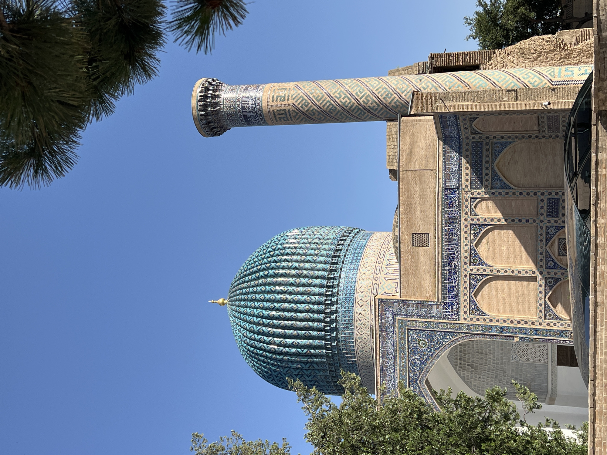 This media file shows a low-angle shot of a section of the Gur-e-Amir Mausoleum in Samarkand, Uzbekistan, set against a clear, bright blue sky. The sun is high, suggesting it is daytime. Green pine branches frame the top left of the image, and leafy green trees obscure the lower portion of the building.
The most prominent architectural features are a fluted turquoise dome topped with a golden finial, and a tall, fluted minaret decorated with intricate geometric patterns and Arabic calligraphy in blue and beige tiles. The main building is constructed of light brown brick and features extensive decorative tiling with blue and white geometric and floral motifs. Arched openings and windows are visible on the facade. A modern black vehicle is partially visible on the right side of the frame, suggesting this is a tourist site.
The overall impression is one of grandeur and historical significance. The bright sunlight casts sharp shadows, highlighting the detailed tilework. There is no specific activity or event taking place; the image captures a static view of the impressive architecture. The visible text appears to be Arabic script, likely inscriptions or decorative elements on the building's facade.