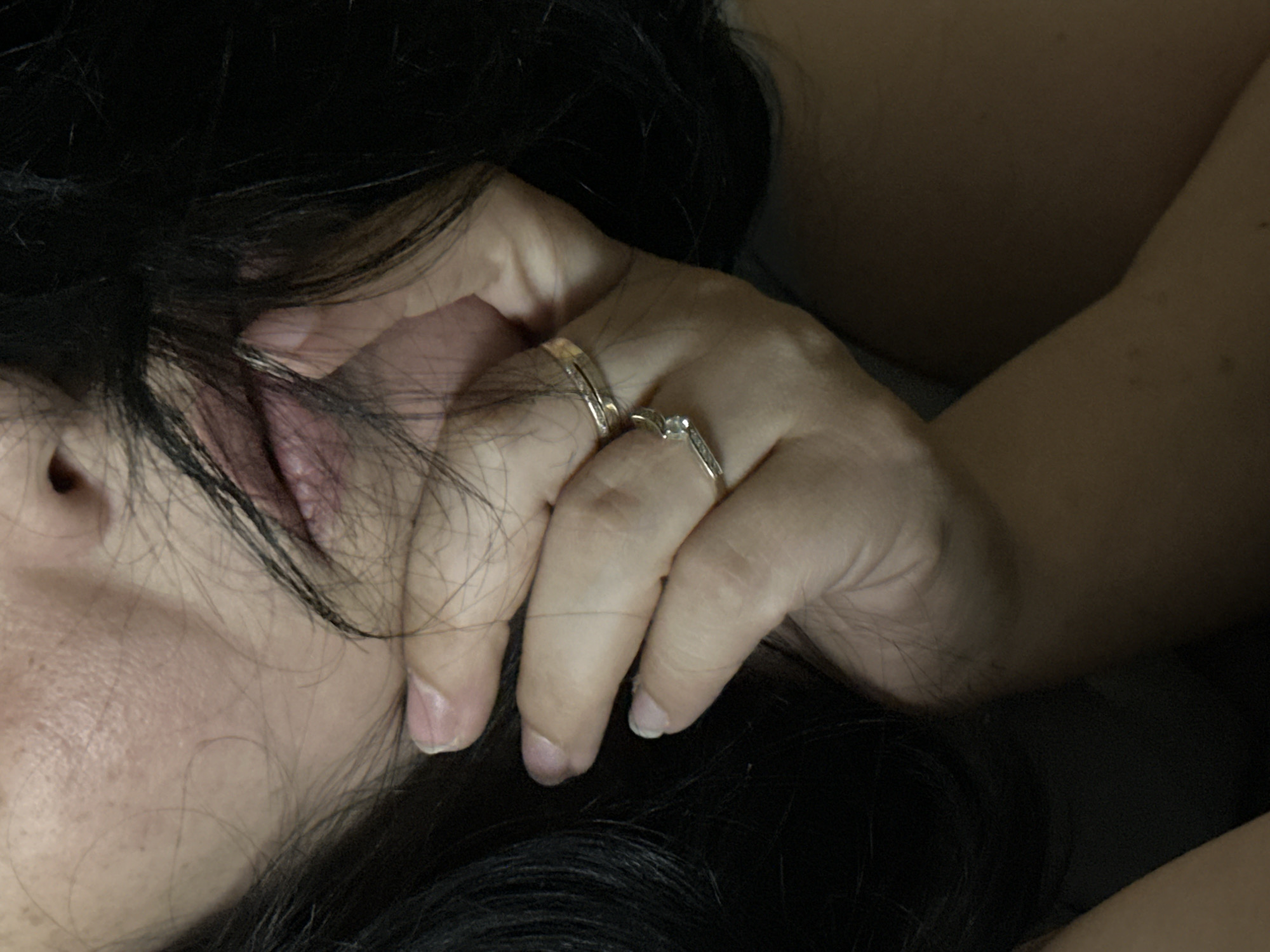 A close-up shot depicts a person resting their head on a dark surface covered with black hair. The person's left cheek, mouth, and nose are visible, revealing details of their skin and facial features. Their hand, adorned with two rings (one gold band and one with a stone setting), is positioned near their face, with fingers partially intertwined with the hair. The overall lighting is subdued, casting shadows and creating a soft, intimate ambiance. The location is Unknown, Unknown.