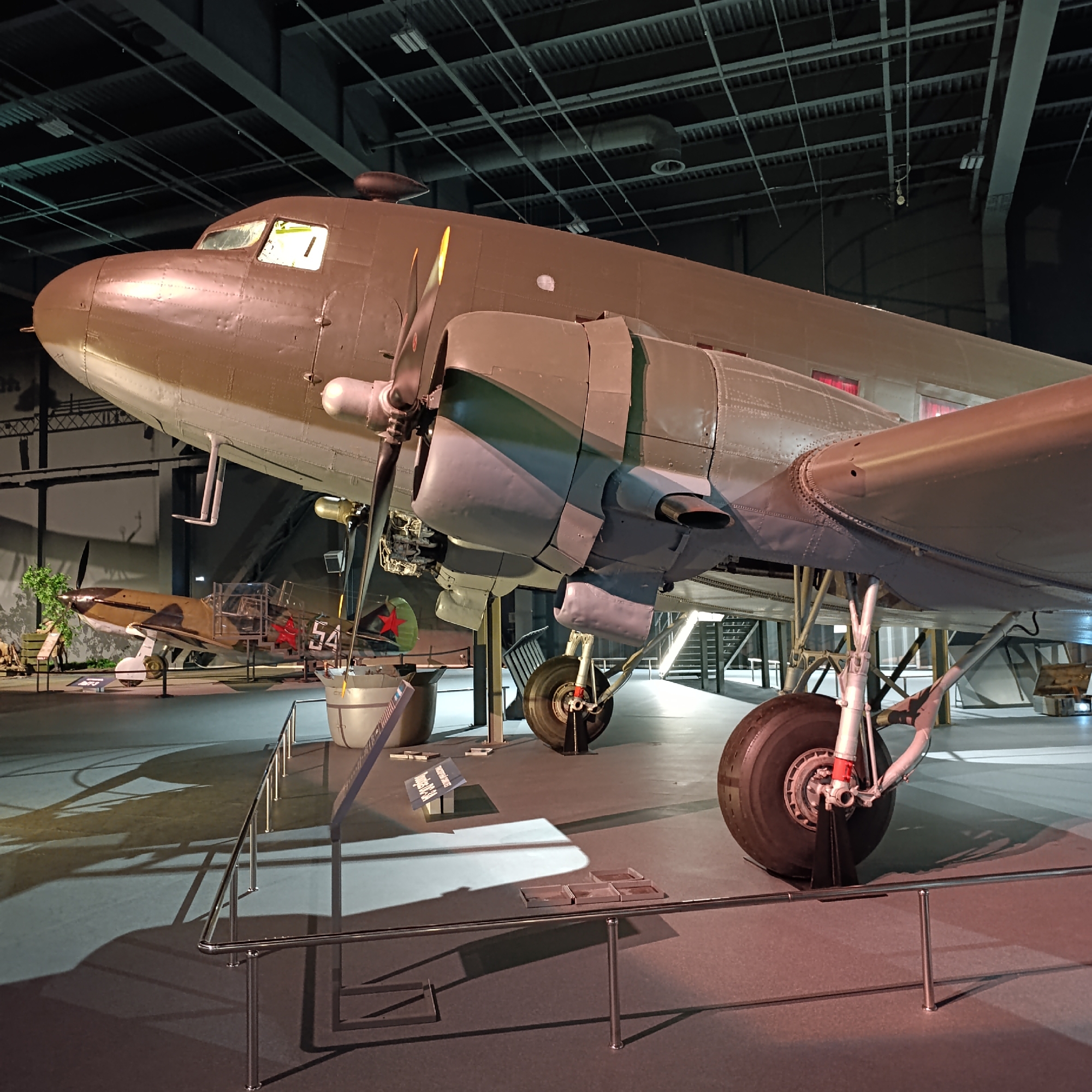 The image captures a detailed view of aircraft on display within a dimly lit indoor museum or exhibition hall, likely located in Verkhnyaya Pyshma, Russia, known for its extensive military museum.
In the foreground, a large, twin-engine propeller-driven transport aircraft dominates the frame. It features an olive drab upper fuselage and lighter underside, with exposed engines, propellers, and prominent landing gear. The aircraft's cockpit windows are visible, and its metal skin shows rivets and minor wear.
Behind and beneath the main aircraft, to the left, a smaller, single-engine fighter plane is partially visible. This fighter sports a brown and green camouflage pattern, with a prominent red star emblem and the number "54" painted on its fuselage, indicative of Soviet-era aviation. Artificial greenery is arranged near this smaller aircraft, suggesting a diorama or historical context.
The scene is set against a dark background with an industrial-style ceiling featuring exposed beams, ventilation ducts, and various lighting fixtures. The floor is dark with reflective areas, and polished metal railings guide visitors around the exhibits. Several information plaques or stands are visible on the floor, one partially readable as "MESSERSCHMITT BF-109", possibly referring to the fighter or another nearby display. No people are visible. The overall impression is one of a meticulously arranged static exhibition, highlighting historical military aviation.