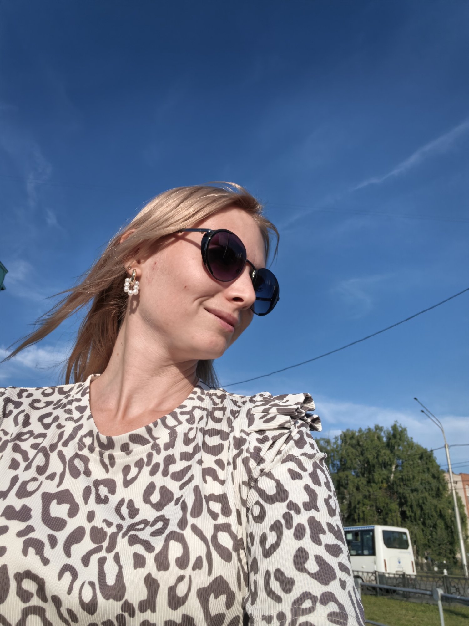 A young woman with blonde hair is photographed outdoors against a bright blue sky with thin clouds. She is wearing large, dark sunglasses and a white top with a leopard print. She has a pearl earring in her left ear. Her hair is blowing to the left, suggesting a breeze. In the background, there is a white bus, trees, a street light, and a building. The scene appears to be on a street in Sterlitamak, Russia, given the visual context of the bus, streetlights, and general urban-like environment.