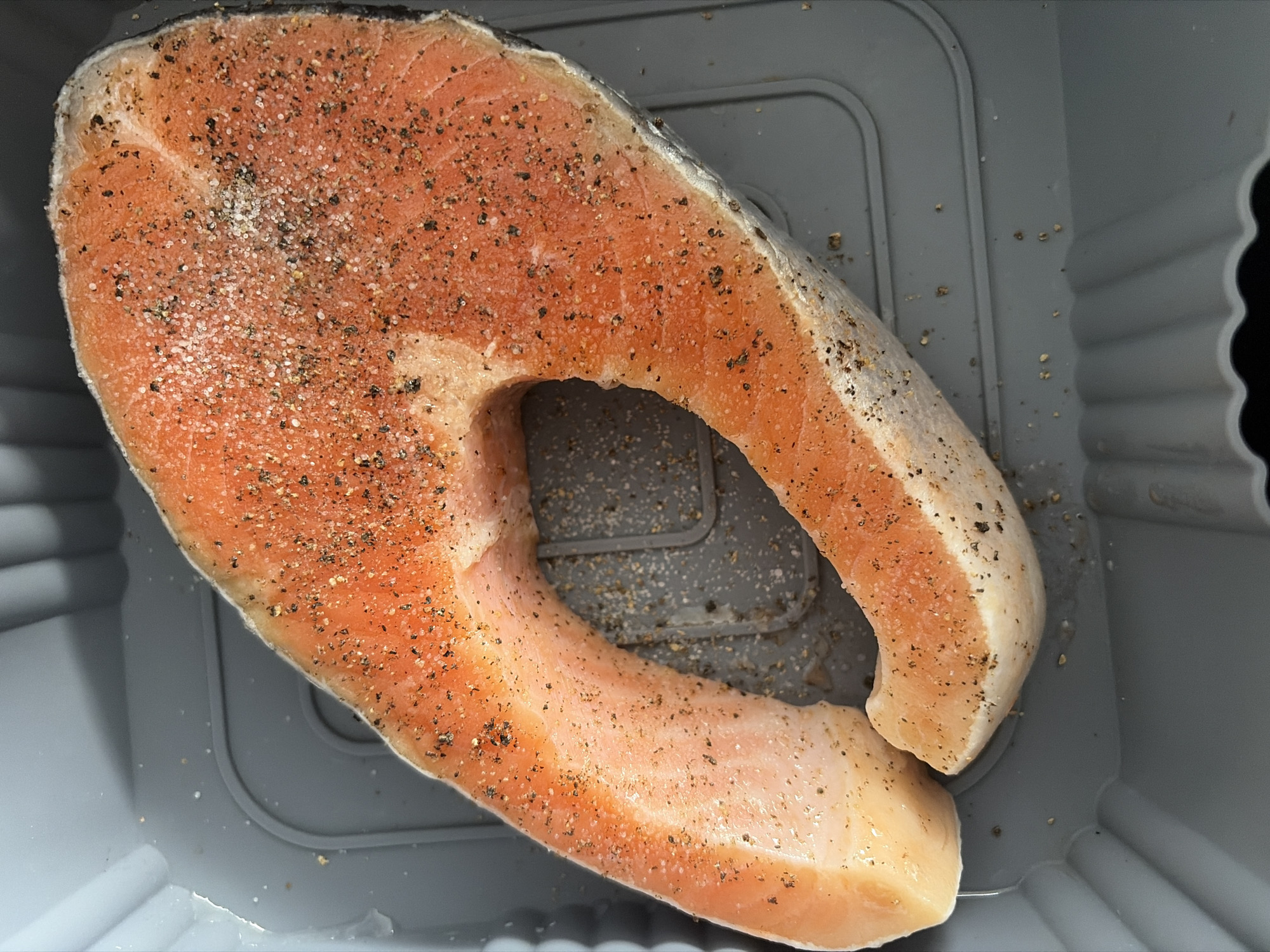 This media file features a close-up, overhead shot of a raw salmon steak seasoned with salt and pepper. The salmon is a vibrant orange-pink color, with visible flakes of seasoning scattered across its surface. It is placed in a gray, rectangular container with ribbed sides. The container appears to be made of plastic, possibly for marinating or storing food. The lighting suggests an indoor setting, as there are no indications of natural light or outdoor elements. The image is focused on the salmon and its preparation, implying an activity related to cooking or meal preparation. No people are visible in the frame. The overall mood is neutral, focusing on the culinary subject matter. The location context provided is Sochi, Russia, but there are no specific visual cues in the image to confirm this. There is no visible text in the media file.