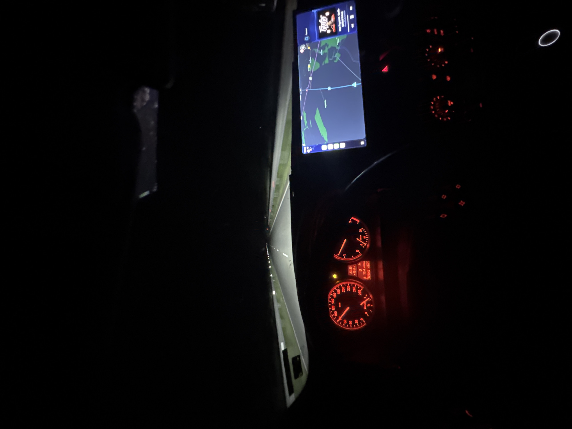 The image depicts the interior of a car at night, likely taken from the driver's perspective. The dashboard and instrument cluster are illuminated in red, displaying speed, RPM, and other vehicle information. A navigation system screen shows a map, indicating the vehicle's location on a road. The road ahead is visible through the windshield, illuminated by the car's headlights.
Given the OCR text "+19.0 D A 10.4 329052 416.1", the coordinates 329052 and 416.1 don't seem to correspond to standard latitude and longitude formats, nor do they immediately suggest a location in Mogila, Bulgaria or elsewhere. The text may represent internal vehicle metrics or non-geographical data.