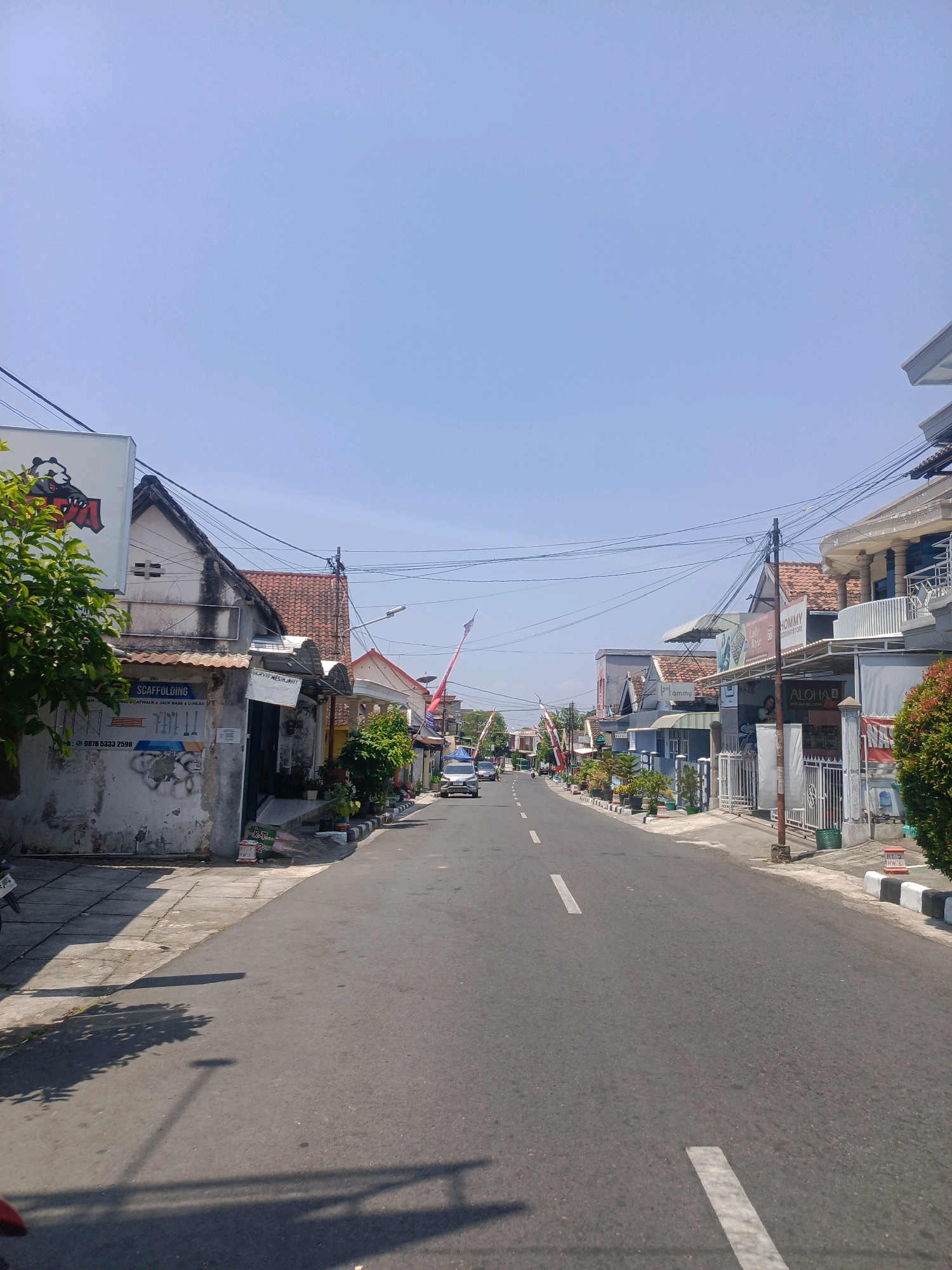 The image depicts a street scene in Kebonagung, Indonesia, under a clear blue sky. The asphalt road stretches into the distance, marked with white lines indicating lanes. On both sides of the road are buildings, some appearing to be shops or small businesses, with awnings and signage visible. Trees and potted plants line the sidewalks. A few cars are present on the road, and utility poles with numerous wires are visible. The architecture suggests a mix of traditional and more modern structures. The overall impression is a quiet, sunny day in a residential or commercial area.