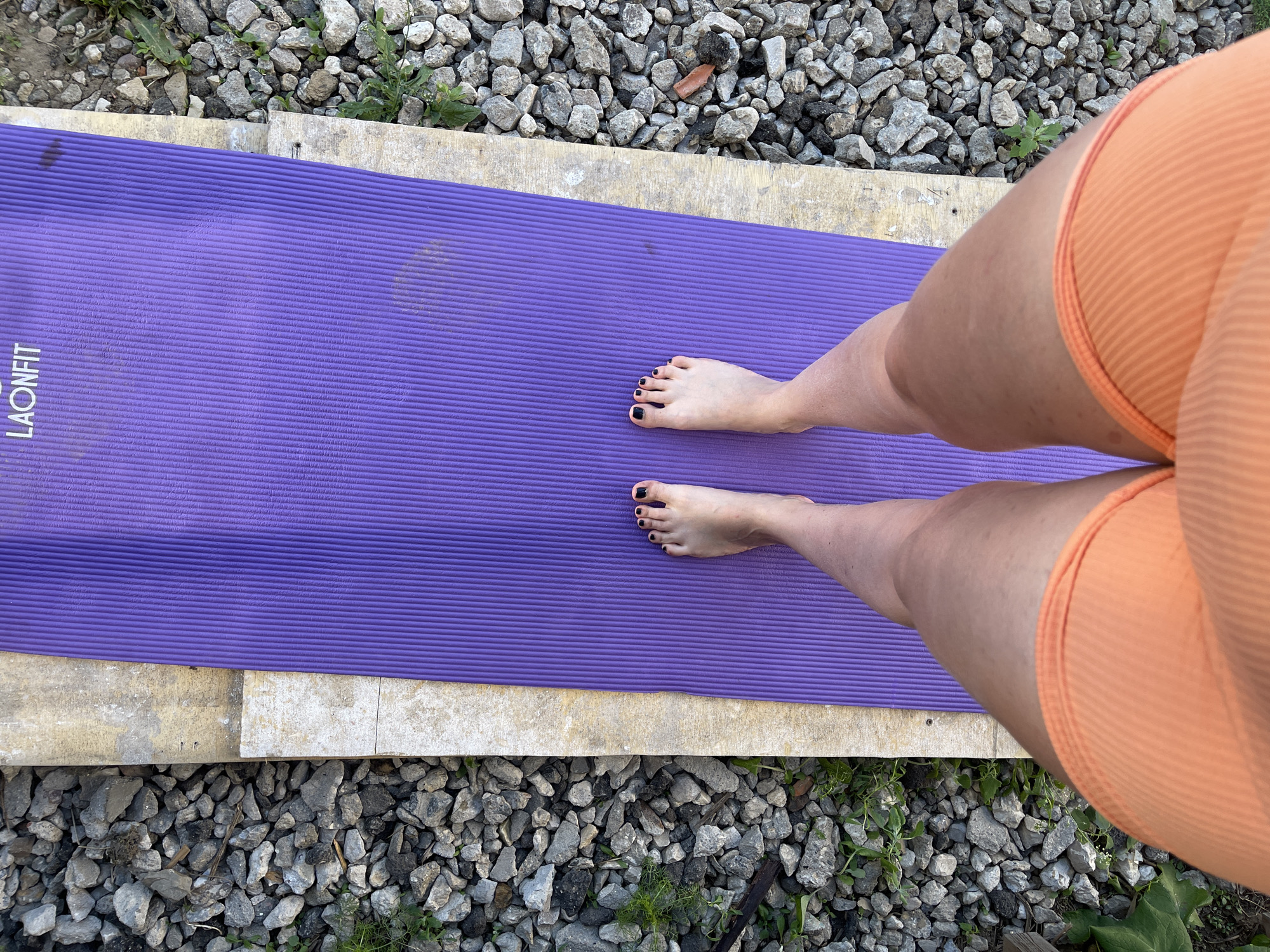 The image shows a person standing on a purple yoga mat outdoors. The person is wearing orange shorts, and their feet are bare with black nail polish. The yoga mat is placed on a concrete surface, and the ground surrounding it is covered with small gray rocks. The environment includes some green plants interspersed among the rocks. The image is a top-down view. There's no indication of the location being in Мосметаллоконструкция Онт, Russia.