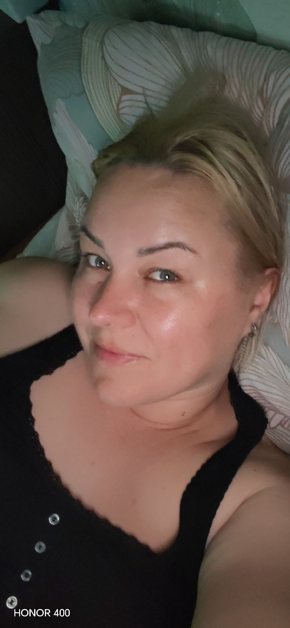 A woman with blonde hair and light eyes is lying on a patterned pillow, wearing a black top with small buttons. The photo was taken in a dimly lit environment. The bottom left corner has the text "HONOR 400". It is impossible to determine the location within the city of Noviy, Russia.