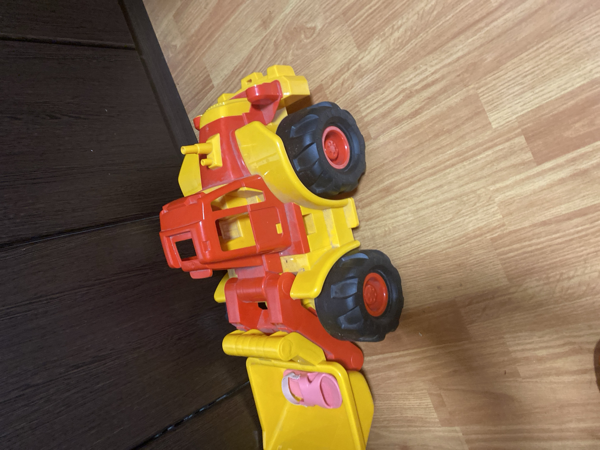 An overhead, slightly angled view shows a brightly colored, red and yellow toy construction vehicle, specifically a loader, resting on a light brown wooden floor. The toy is positioned diagonally, with its front end and bucket to the left of the frame and its rear end to the right. The loader has large, black, treaded wheels with red centers. A pink child's sandal is placed inside the yellow bucket of the toy. The background consists of a dark brown wooden wall with vertical paneling. The scene is indoors and appears to be in a domestic setting, though the specific location within a city is unknown.