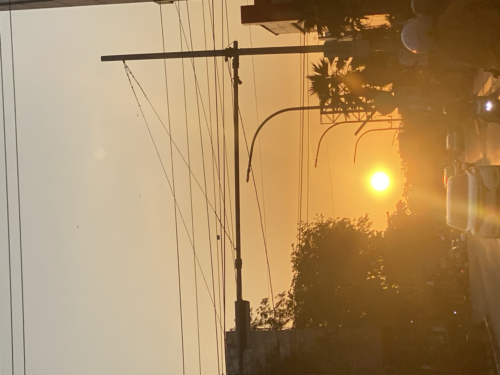 This is a photograph taken in Banjarmasin, Indonesia, during sunset. The sun is a bright, glowing orb near the center of the frame, casting a warm, orange hue across the sky and the street scene below. The sky is a soft gradient of yellow and orange. Overhead, a network of power lines crisscrosses the frame, supported by poles.
On the left side of the image, a section of a building with a concrete structure and some foliage from a tree is visible. The street is lined with trees, some of which are silhouetted against the bright sky. There are several vehicles on the road, including cars and motorcycles. Some people are visible on motorcycles, wearing helmets. The overall atmosphere is one of an urban environment at dusk.
