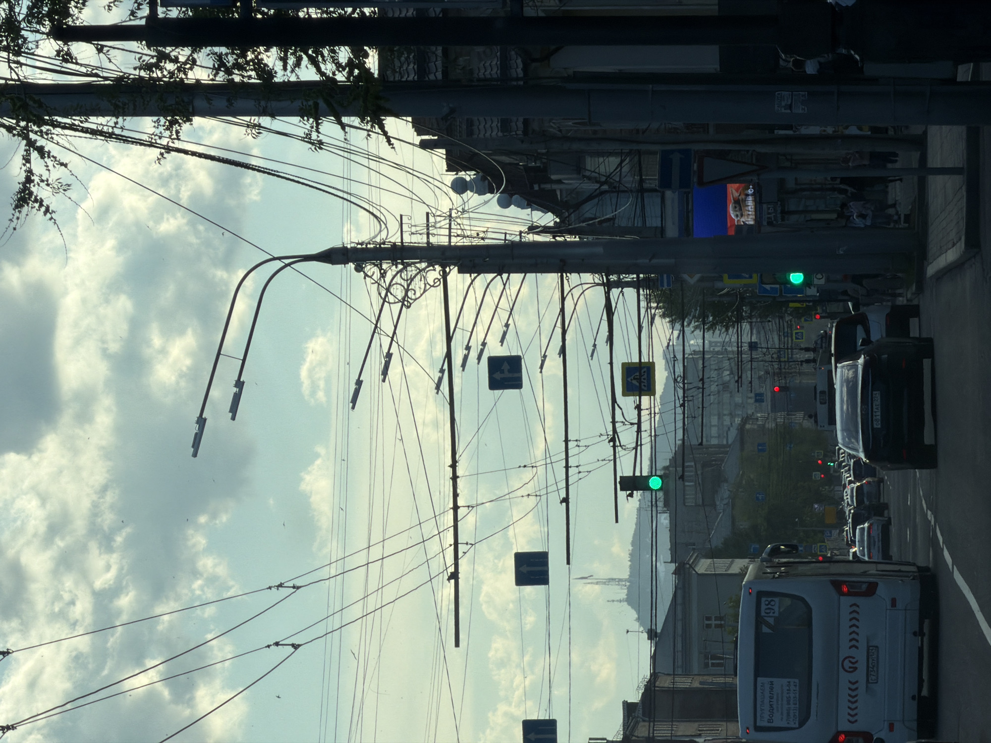 The image shows a street scene in Krasnoyarsk, Russia. Looking down the street, several cars and a white bus are driving in the right lane. Above the vehicles, utility poles with overhead wires stretch across the sky. Traffic lights are visible, with some showing green and others red. A few street signs are attached to the utility poles. Buildings line both sides of the street. Text is visible on the back of the bus, including the numbers "198" and a phone number. The sky is cloudy and blue.