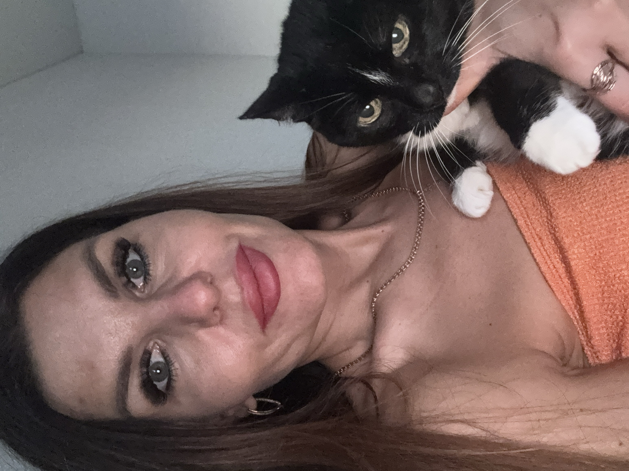 A woman with shoulder-length brown hair and fair skin is holding a black and white cat close to her face. The woman is wearing a gold necklace and earrings, and has red lipstick on. The cat is looking at the camera with green eyes. The background appears to be a light-colored wall. The specific location in the city of Chaikovsky, Russia, cannot be determined from the image.