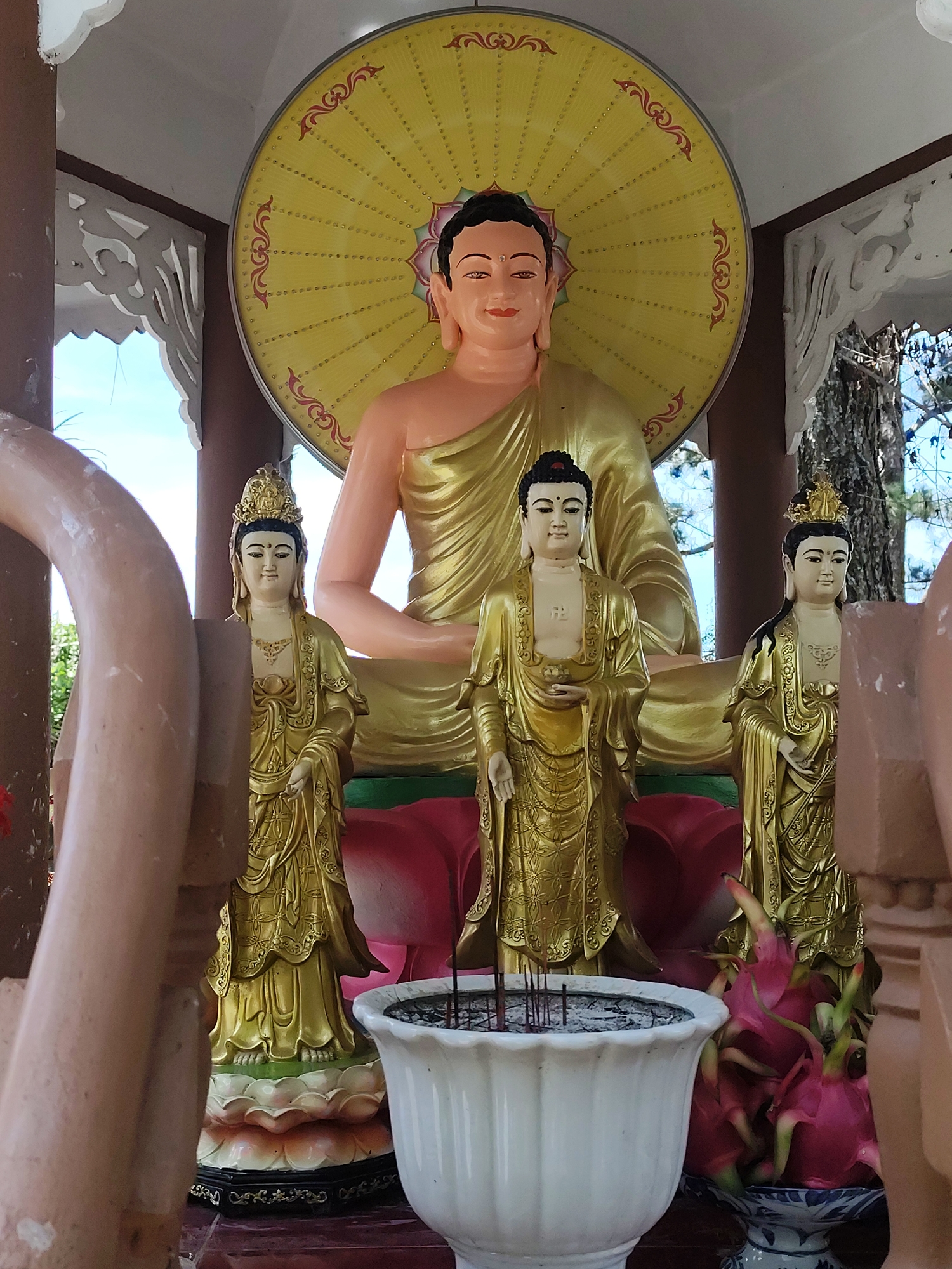 The image shows a Buddha statue at a temple in Xuân Trường - Đà Lạt, Vietnam. The statue features a central seated Buddha figure with a yellow halo behind him. Two smaller, golden-robed figures stand on either side of a central, shorter figure also in golden robes, all displayed atop a multi-tiered lotus base. An incense holder filled with incense sticks sits in front of the figures. Dragon fruit is visible in the bottom right corner. The statues are housed in a partially enclosed structure with decorative white trim and dark wooden posts. Portions of other architectural elements are seen in the foreground, partially obscuring the view of the statue.