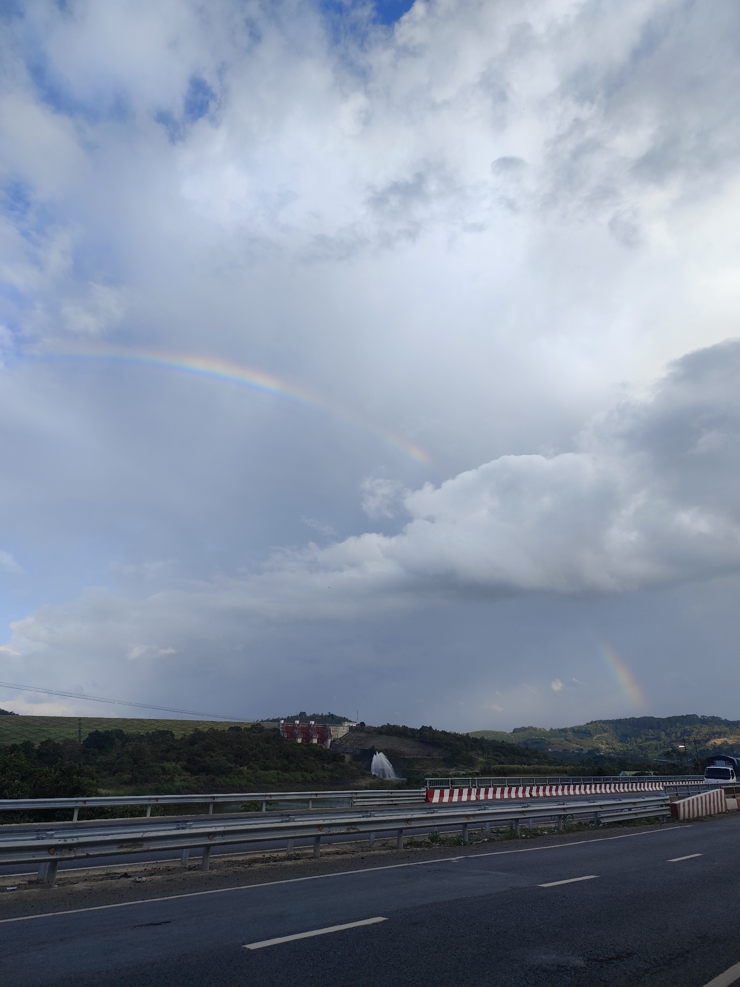 The image shows a dual rainbow arcing across a cloudy sky over a landscape that includes a highway, guardrails, rolling hills, and structures that appear to be related to a dam or water management facility. In the middle distance, on a hillside, are several red-roofed buildings and a large white structure with water gushing out, possibly a spillway or fountain. The highway in the foreground is asphalt with white lane markings, bordered by metal guardrails and concrete barriers with red and white stripes. The sky is filled with dramatic, grey clouds, with patches of blue visible. The overall scene suggests a natural event (the rainbows) occurring over an engineered landscape. The location is identified as the city of Phú An, Vietnam.