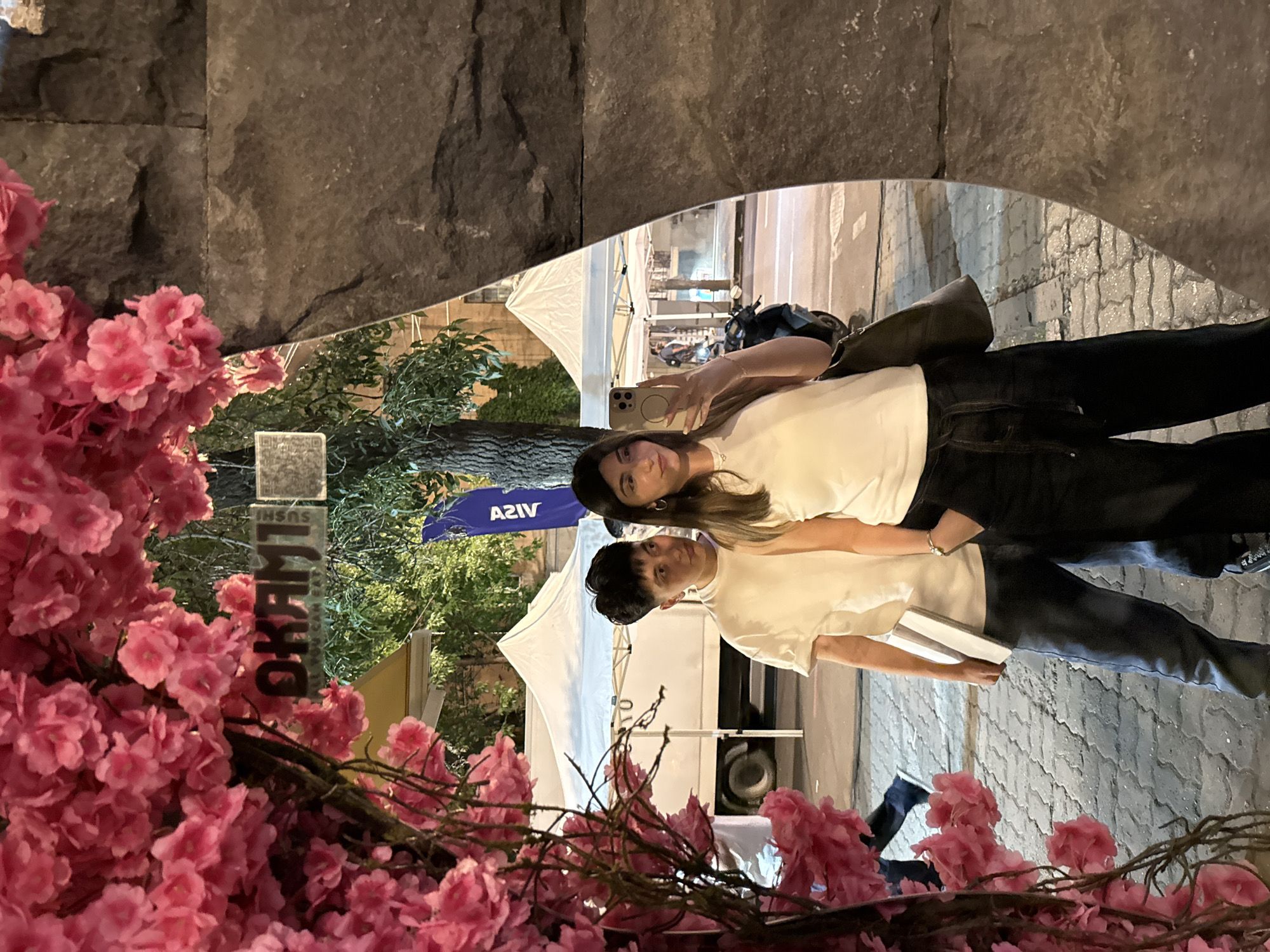 This media file captures two young individuals framed by a decorative archway made of dark, textured stones and adorned with abundant artificial pink cherry blossoms. The scene is set outdoors at night or late evening, as indicated by the ambient lighting, illuminated tents, and overall dimness.
The primary subjects are a young woman and a young man. The woman, with long dark hair, wears a white t-shirt and dark pants. She holds a silver smartphone up, seemingly taking a selfie or video, and offers a soft smile to the camera. A small earring and a bracelet are visible. Beside her, the man, with short dark hair, also wears a white t-shirt and dark pants, holding a stack of white books or a similar rectangular object. He looks towards the camera with a neutral expression.
In the background, beyond the subjects, white event tents are visible, along with green trees and a partially obscured parked scooter or motorcycle. The ground is paved with stones or bricks. The composition suggests a public outdoor event, market, or festival in an urban setting.
Notable details include the vibrant pink artificial blossoms, which create a striking contrast with the dark stone frame. The overall atmosphere appears relaxed.
Visible text includes "OKAMI SUSHI BAR" on a vertical sign, partially hidden by the blossoms, and the "VISA" logo on a blue banner or flag in the background. The location context provided is Yerevan, Armenia.