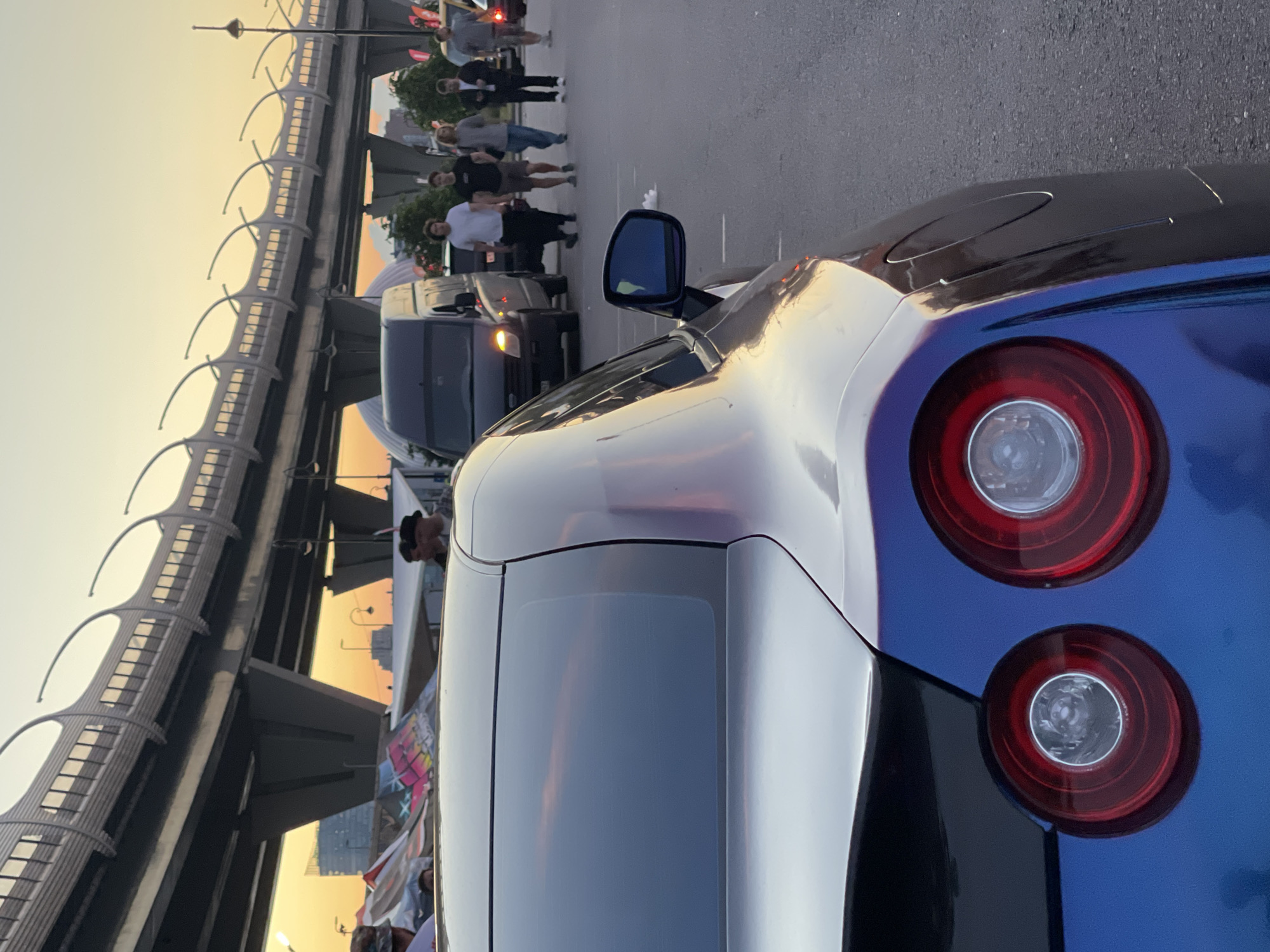 In an outdoor urban setting at dusk, a blue and silver sports car with distinctive round taillights is parked in the foreground, partially obscuring the view. Behind the car, a group of people walk on a paved surface. A dark-colored van is also present. An elevated highway structure with curved supports stretches across the background, illuminated by the warm glow of the setting sun. The sky displays vibrant hues of orange and yellow. A few buildings and lampposts are visible in the distance. The scene appears to be a public space, possibly a parking lot or street near an event.