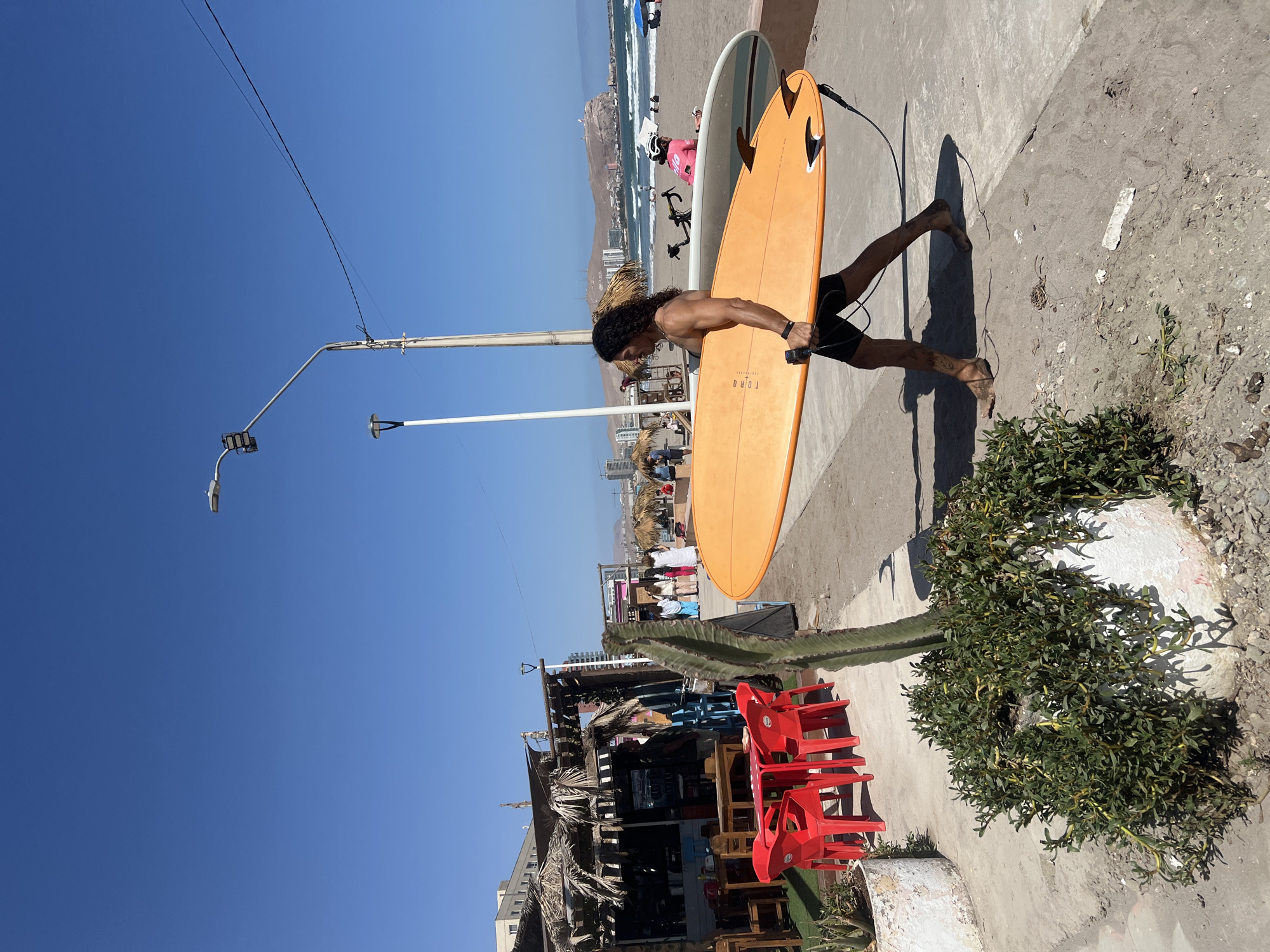 A full-length, medium shot photograph captures a surfer carrying two surfboards along a concrete walkway on a sunny day in Arica, Chile. The man, barefoot and wearing dark shorts, is seen from the side as he walks from right to left across the frame. He holds an orange surfboard under his right arm, with a larger, light-colored surfboard resting against it.
In the background, the beach is visible, with people, umbrellas, and the ocean stretching towards the horizon. Buildings and hills line the coast in the distance. To the left of the surfer, there's a street scene with a thatched-roof structure, red plastic chairs, and various details suggesting a local beachside establishment. Above, streetlights and power lines cut across a clear, blue sky.
To the right of the surfer, the concrete walkway has some debris and vegetation, including a patch of green plants growing around a white planter. The lighting is bright and harsh, creating strong shadows. The overall scene suggests a typical day at a surf spot in Arica.