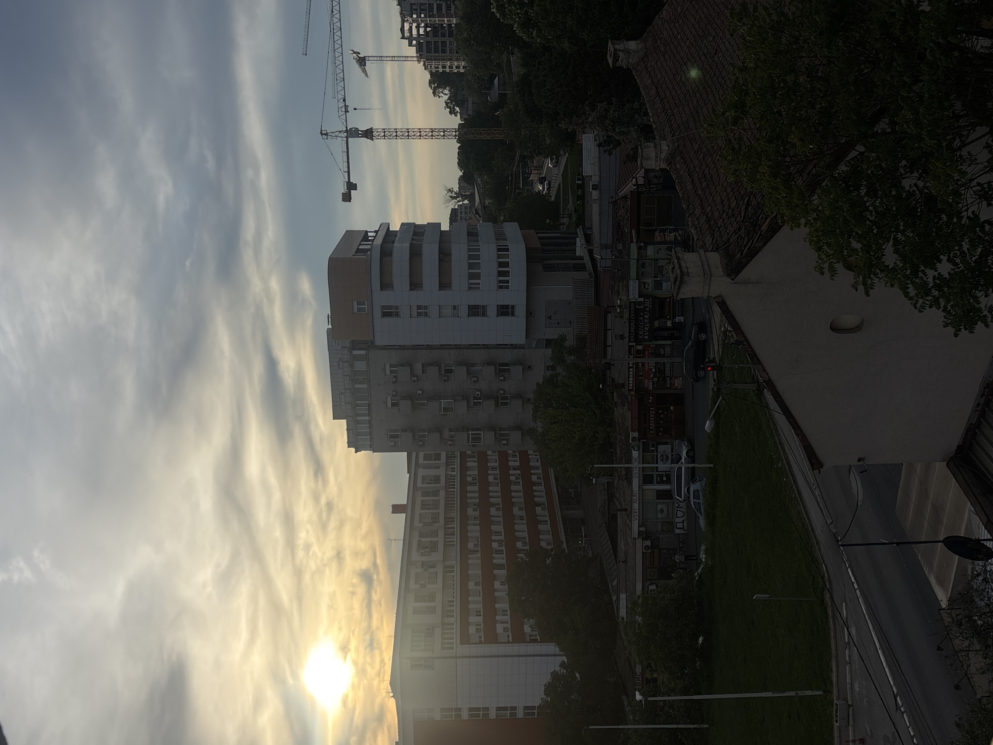 The image shows an urban landscape in Belgrade, Serbia. The scene includes a modern apartment building, a construction site with cranes, a street with commercial establishments, and older residential buildings. The sky is cloudy with the sun partially obscured. There are no visible people. The perspective is from an elevated vantage point, possibly a building or apartment. The overall environment suggests a mix of ongoing development and existing infrastructure.