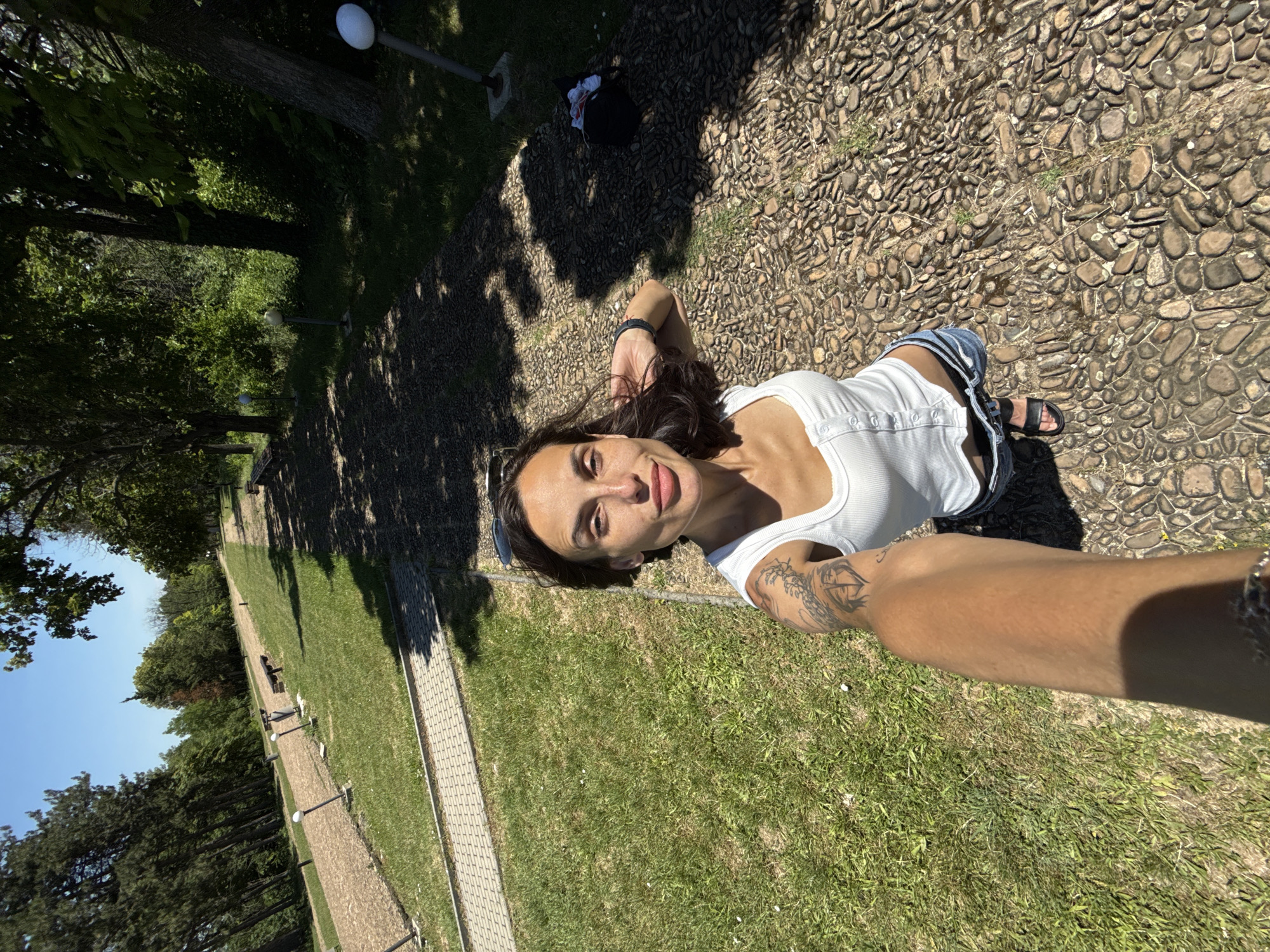 In Pleven, Bulgaria, a woman stands on a pebble pathway next to a grassy area. She is taking a selfie, holding the camera with her right arm, which is extended. The woman is wearing a white tank top and denim shorts, with black sandals. She has tattoos on her left arm. Behind her, the pathway curves through a park-like setting with trees and benches. The sky is clear and blue, indicating a sunny day.