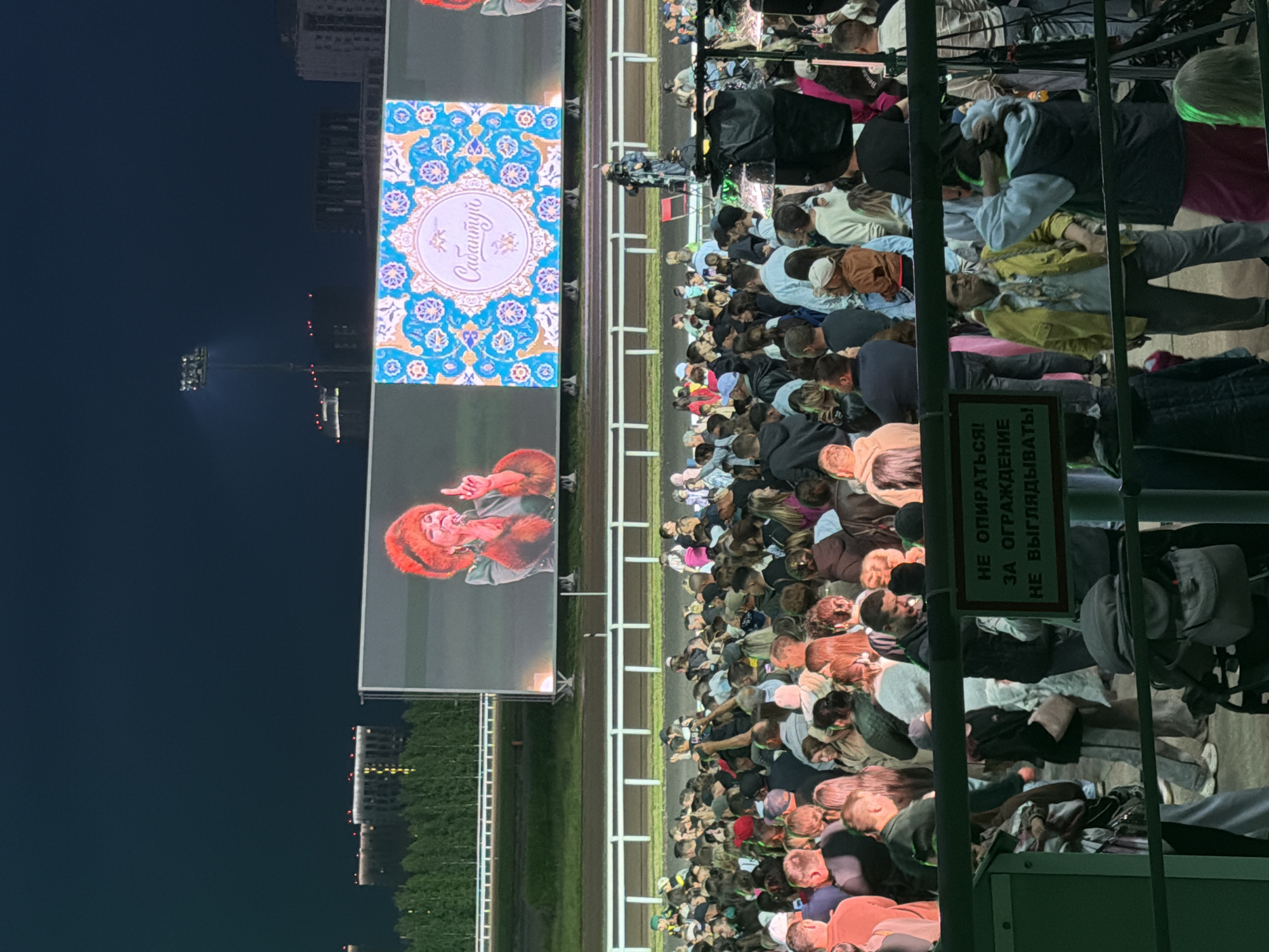 An outdoor evening concert is taking place, with a large crowd gathered in front of a massive LED screen displaying a performer and decorative graphics. The performer, a woman wearing a red fur hat and coat, is shown singing into a microphone. The screen also features a blue and gold ornamental pattern with the word "Сабантуй" in cursive script. The audience appears diverse in age and gender, with many people holding up their phones to record the event. The environment includes a race track in the background, illuminated by stadium lights, and residential buildings in the distance. A sign in Russian is visible in the foreground, advising people not to lean on the fence. The scene suggests a festive and energetic atmosphere.
