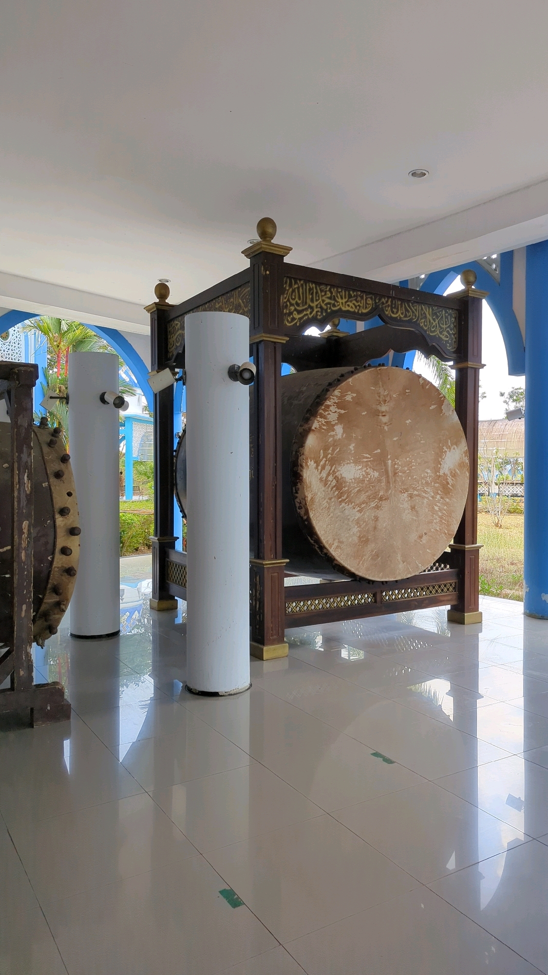 The image depicts a large, traditional drum, likely a *bedug*, displayed on a wooden stand decorated with intricate Arabic calligraphy. The drum is positioned outdoors, under a covered walkway with a tiled floor that reflects the surrounding light and structures. Two white cylindrical pillars are visible in the foreground, and to the left, another drum-like object with studded edges can be partially seen. The background reveals lush greenery, palm trees, and blue architectural elements, suggesting a tropical or subtropical environment, possibly associated with a place of worship or a cultural venue in Gunungtinggi, Indonesia. The lighting indicates it is daytime.