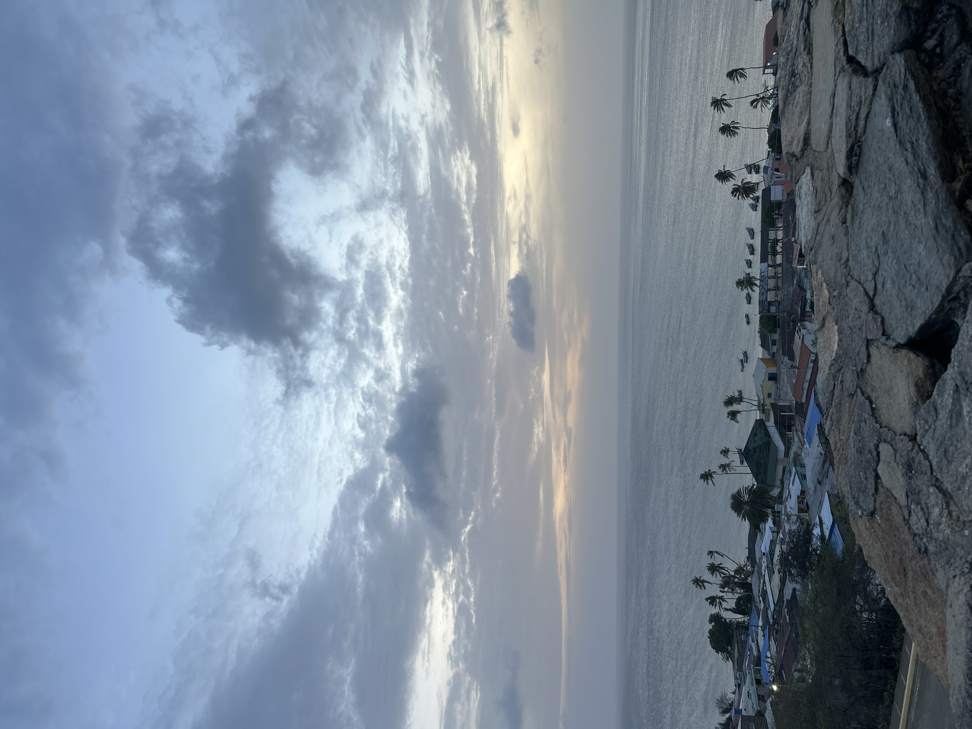 The image captures a coastal landscape in Juan Griego, Venezuela, during an overcast dawn or dusk. The upper two-thirds of the frame are dominated by a cloudy sky, transitioning from lighter blue at the top to various shades of grey, blue-grey, and off-white. A subtle band of pale yellow and orange light is visible on the horizon, obscured by clouds, indicating a low sun angle.
The sea occupies the middle ground, appearing calm with a muted grey-blue tone and minimal surface disturbance. Along the coastline, a cluster of residential structures forms a town, characterized by buildings with diverse roof colors, including blue, green, grey, and reddish-brown. Numerous palm trees, some appearing windswept, are interspersed among the buildings and line the shore. Several small boats are visible moored in the water near the town's edge.
The immediate foreground features a textured, grey stone wall or embankment, indicating the elevated perspective from which the photograph was taken. No individuals are explicitly depicted in the scene.